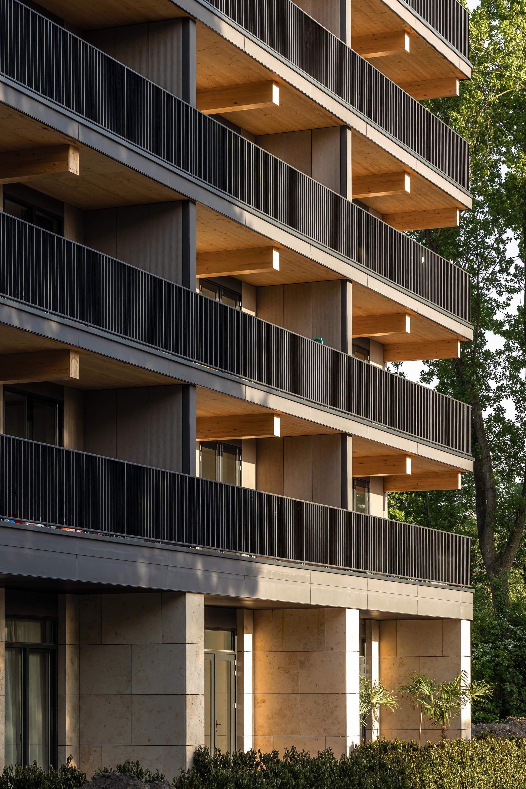 Multi-story facade showing timber balcony soffits and concrete base with planted beds at ground level