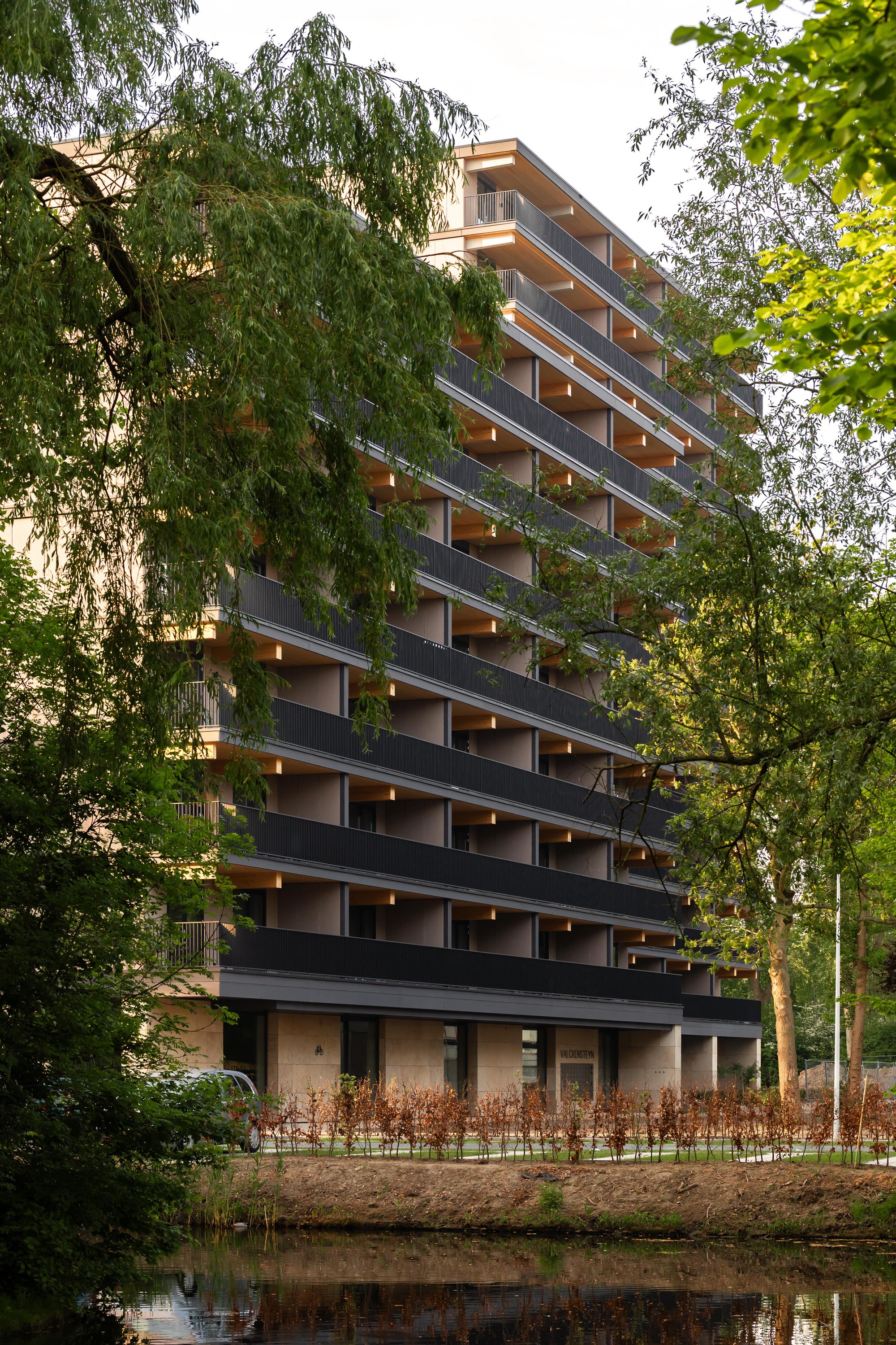 Tower facade with cantilevered balconies reflected in the canal at dusk through foliage