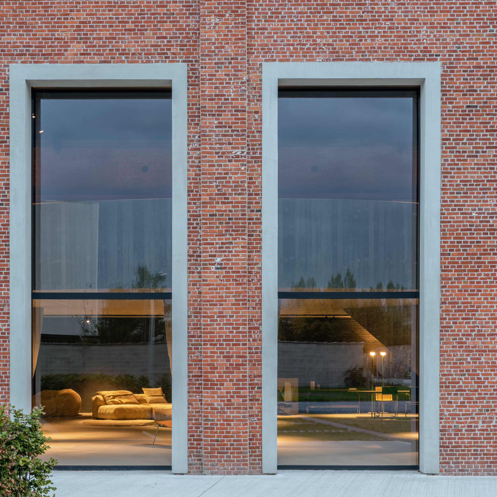 Two tall glazed openings framed in pale plaster reveal interior lounge furniture at dusk