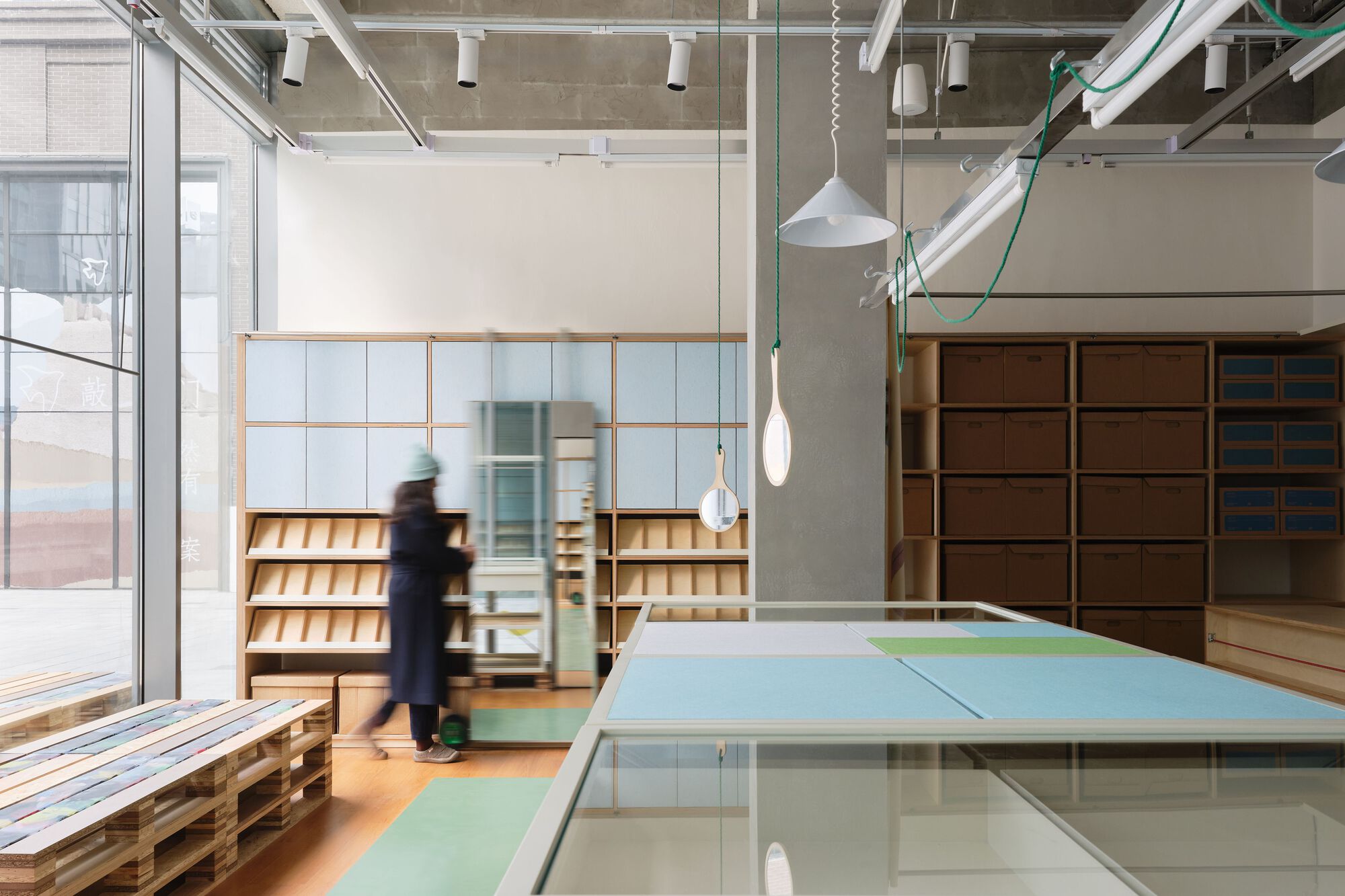 Interior retail space with exposed concrete ceiling and a person passing glass partitions near timber shelving