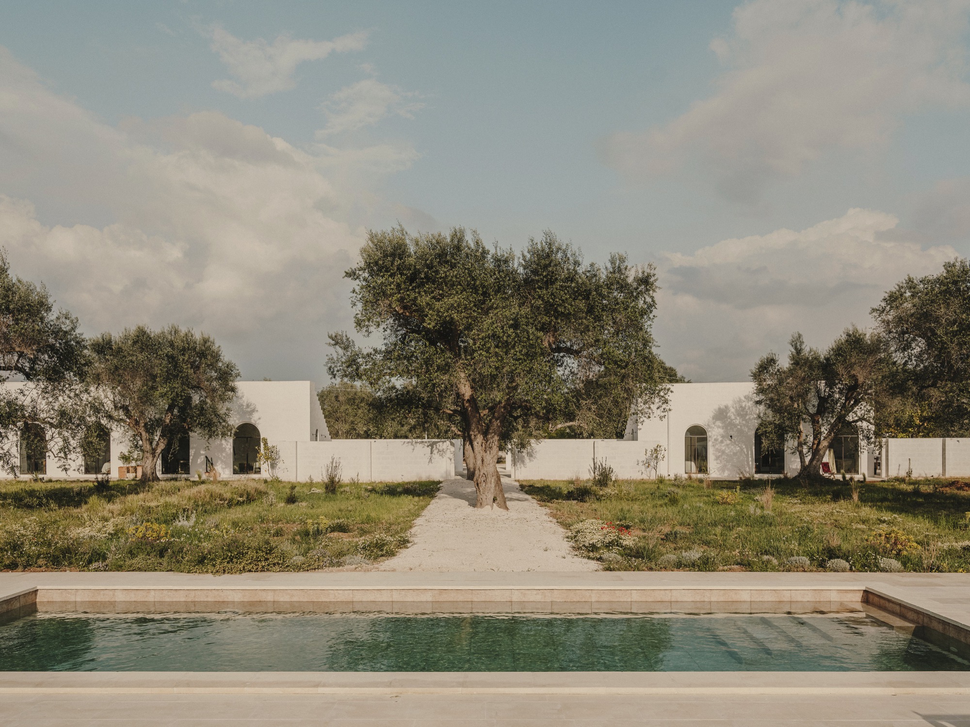 View across the swimming pool toward white pavilions with arched openings framed by olive trees