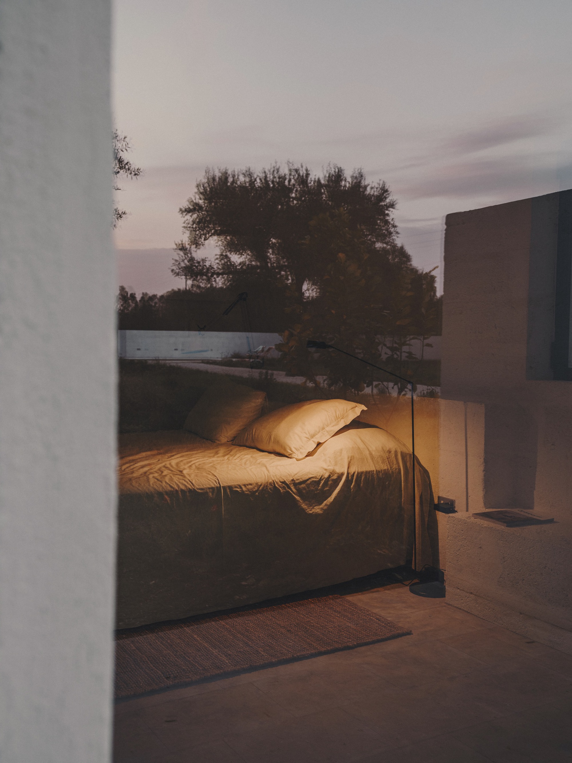 Bedroom with unmade linen bedding glimpsed through doorway at dusk with trees beyond glass wall