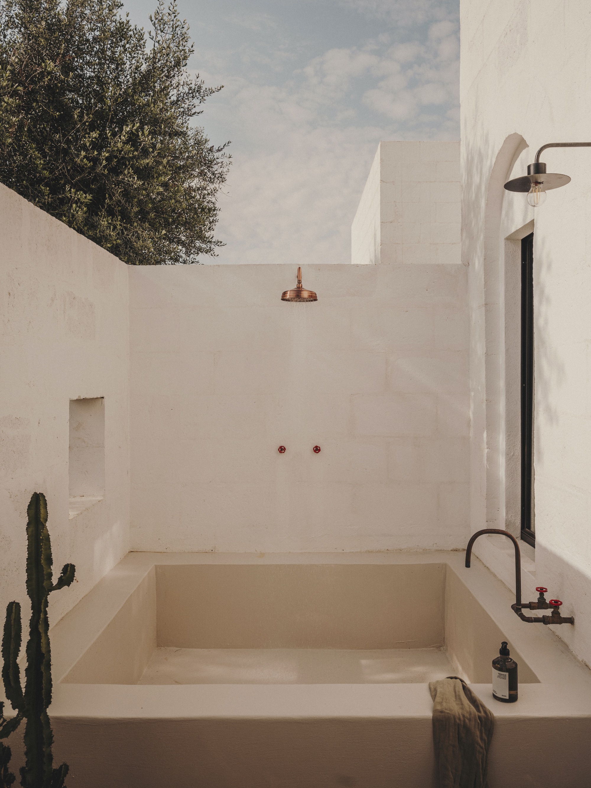 Outdoor bathing area with sunken tub, copper rain showerhead, and a cactus beside white plastered walls