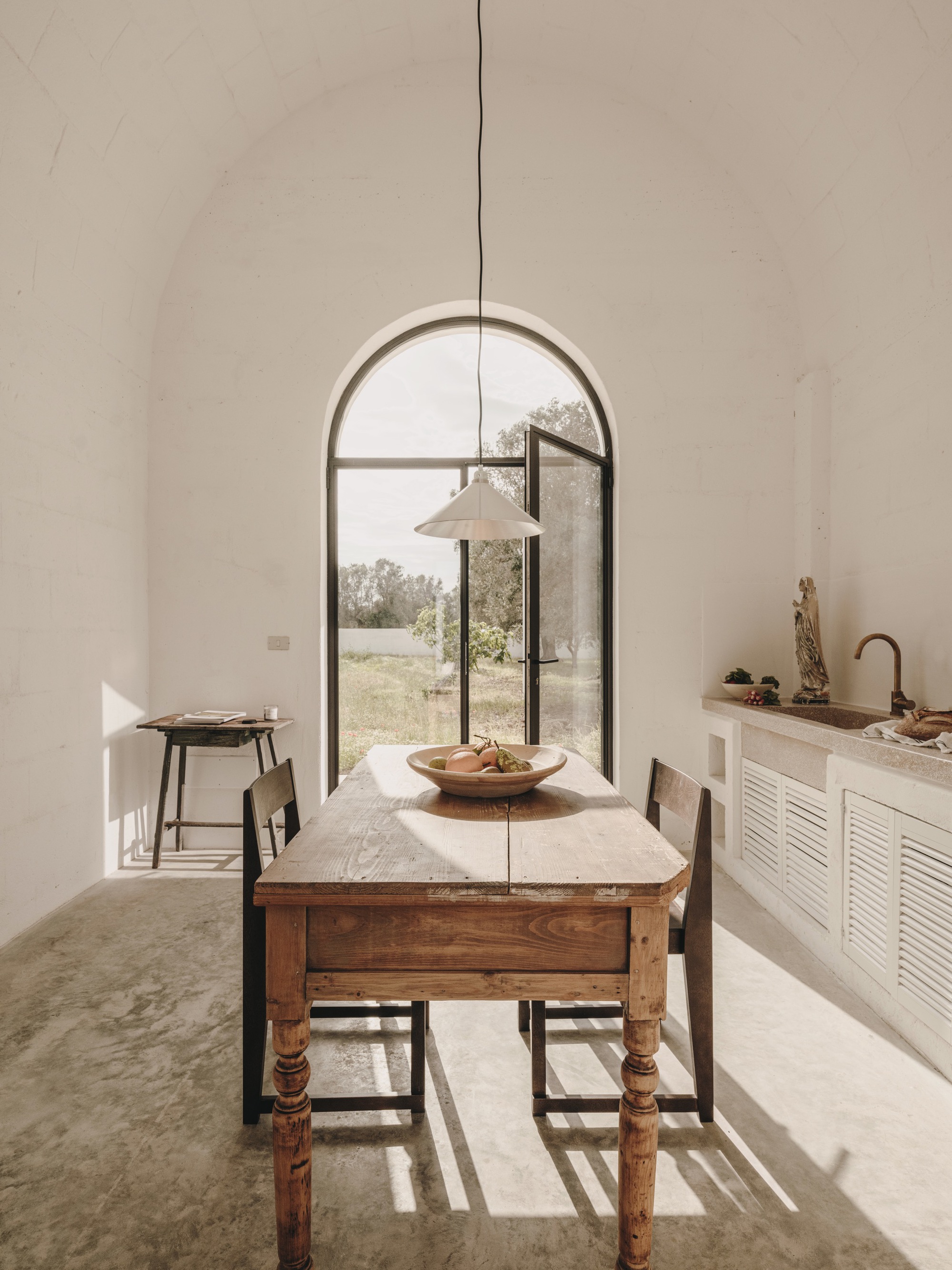Dining area with wooden farmhouse table, pendant lamp, and arched glazed door overlooking an olive grove