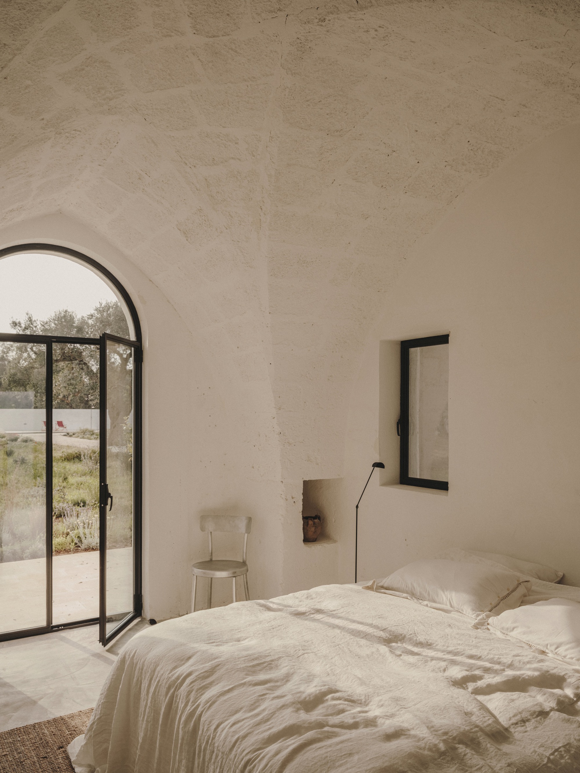 Bedroom with arched steel-framed glass door opening to garden with olive trees beyond