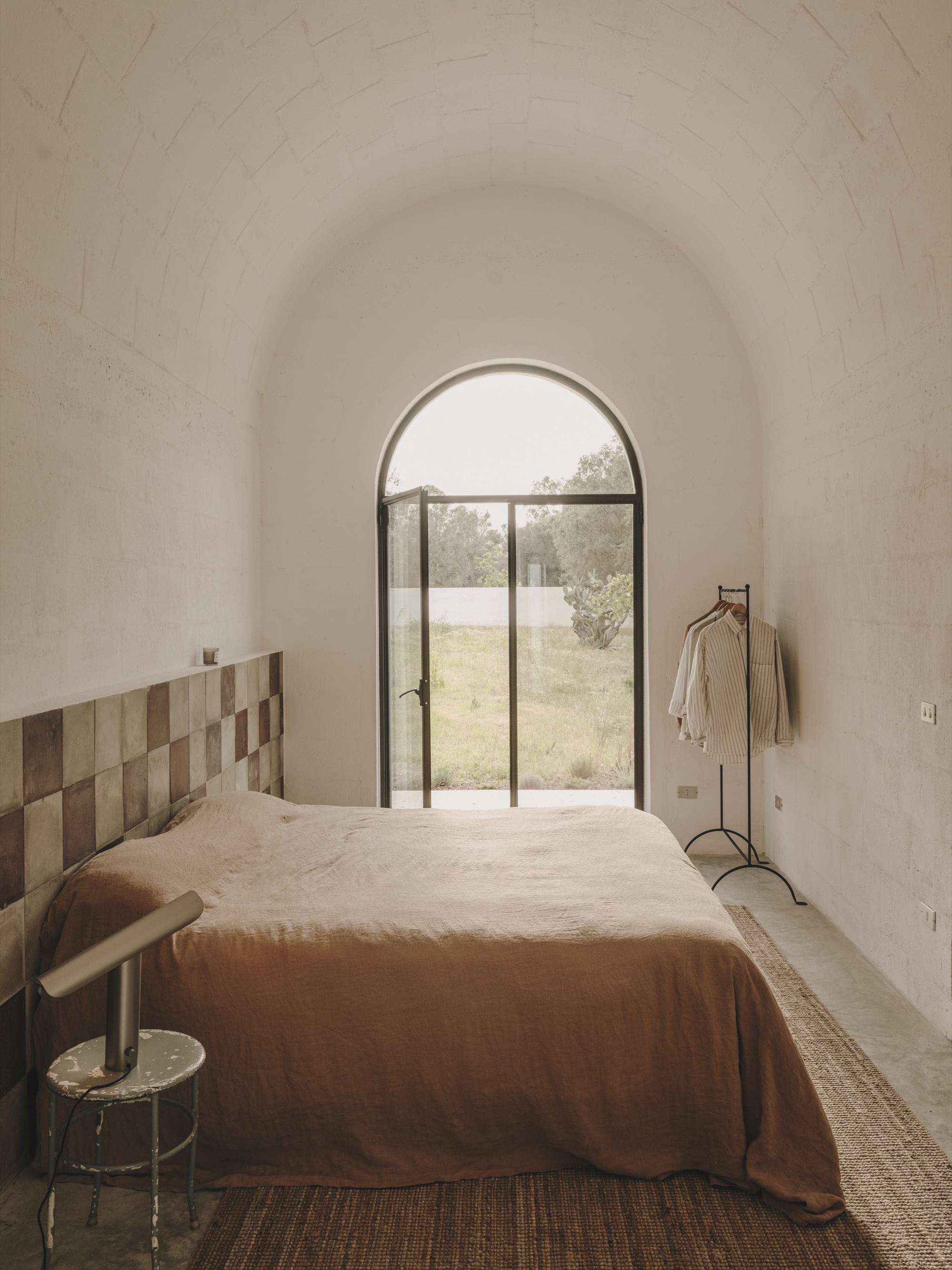 Bedroom with patchwork timber headboard and arched steel door framing view of olive grove