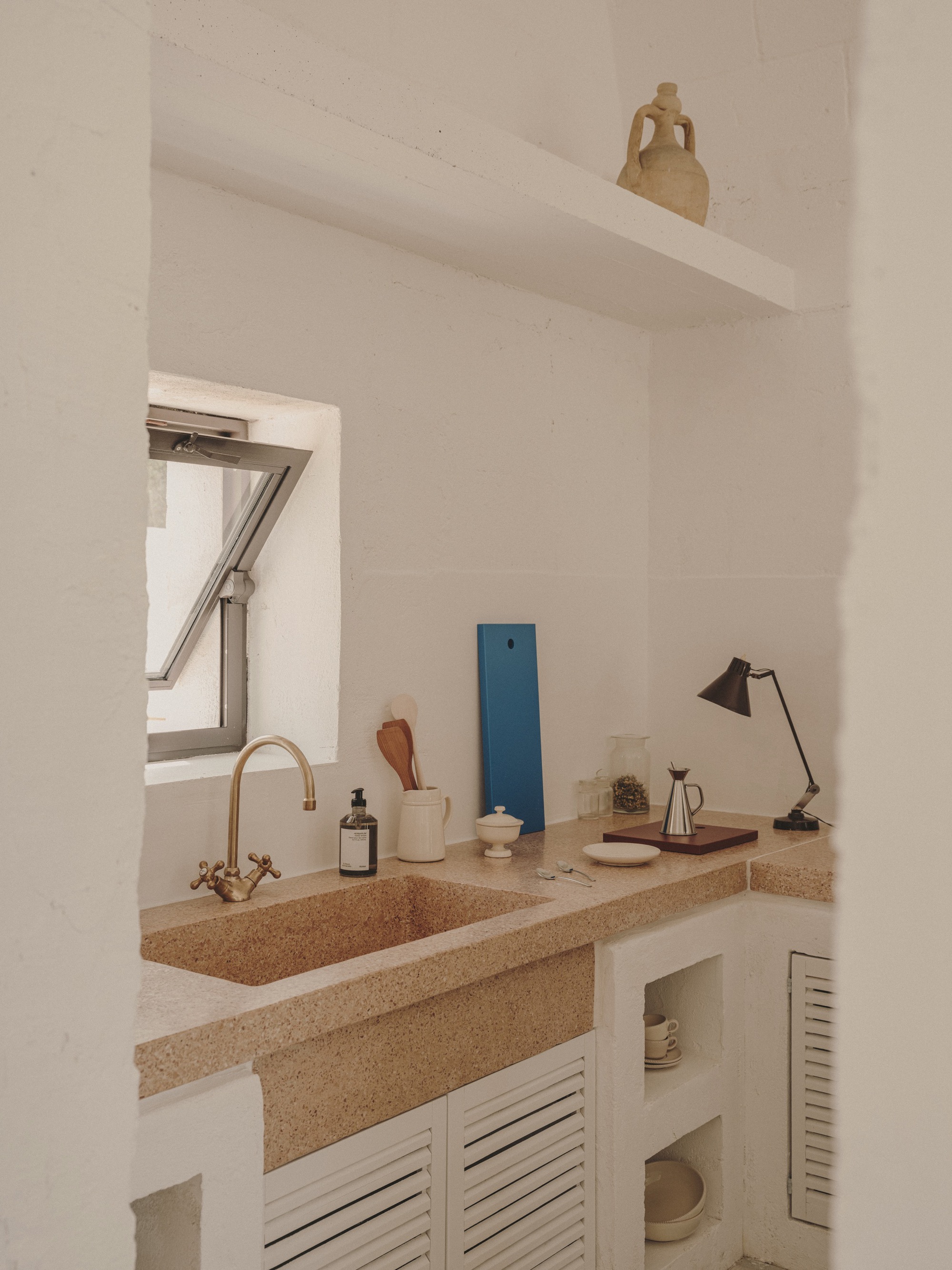 Kitchen counter with terrazzo surface, brass faucet and louvered cabinet doors below skylight