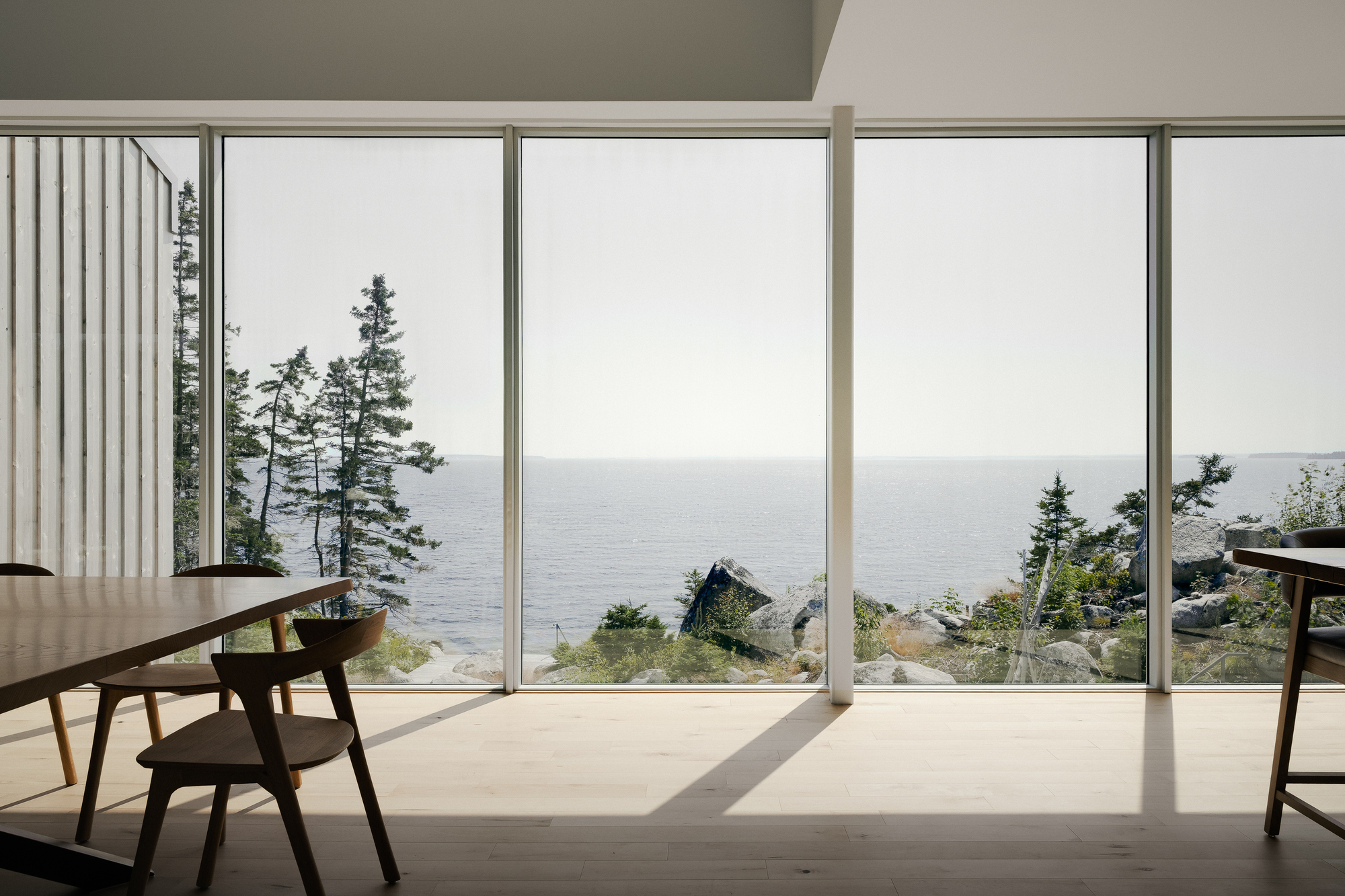 Floor-to-ceiling glass wall framing distant water and coastal pines with timber chair in sunlight