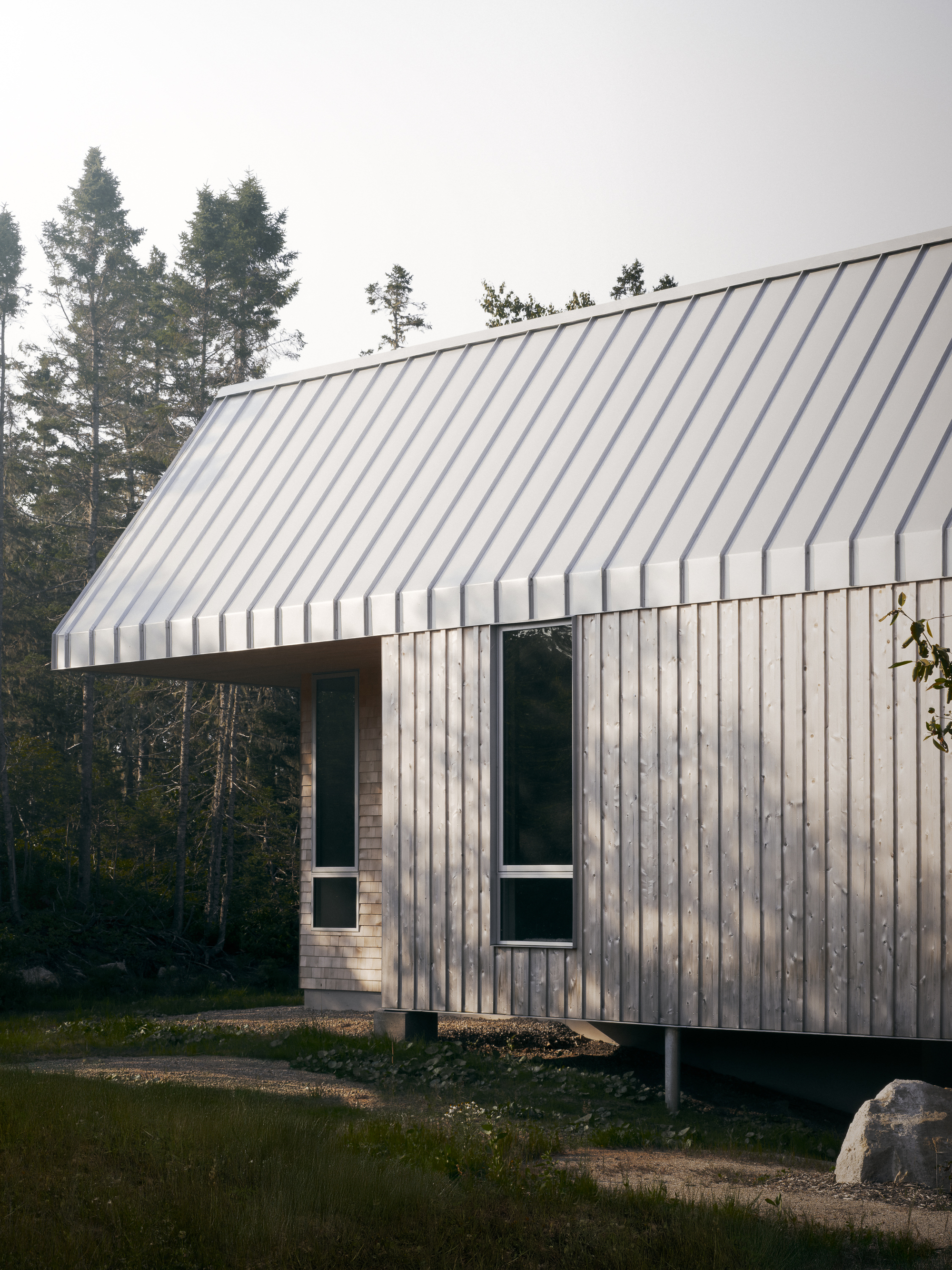 Gable-roofed volume with vertical timber siding elevated on concrete piers among evergreens