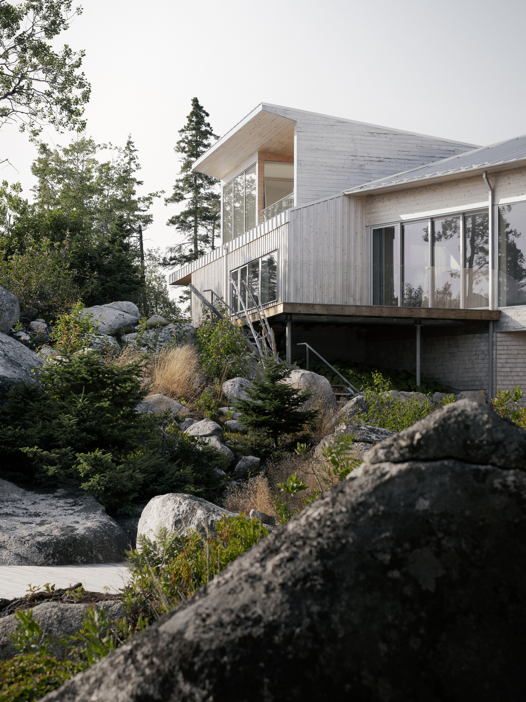 Two-story glazed corner stairwell connecting timber volumes above a rocky planted slope