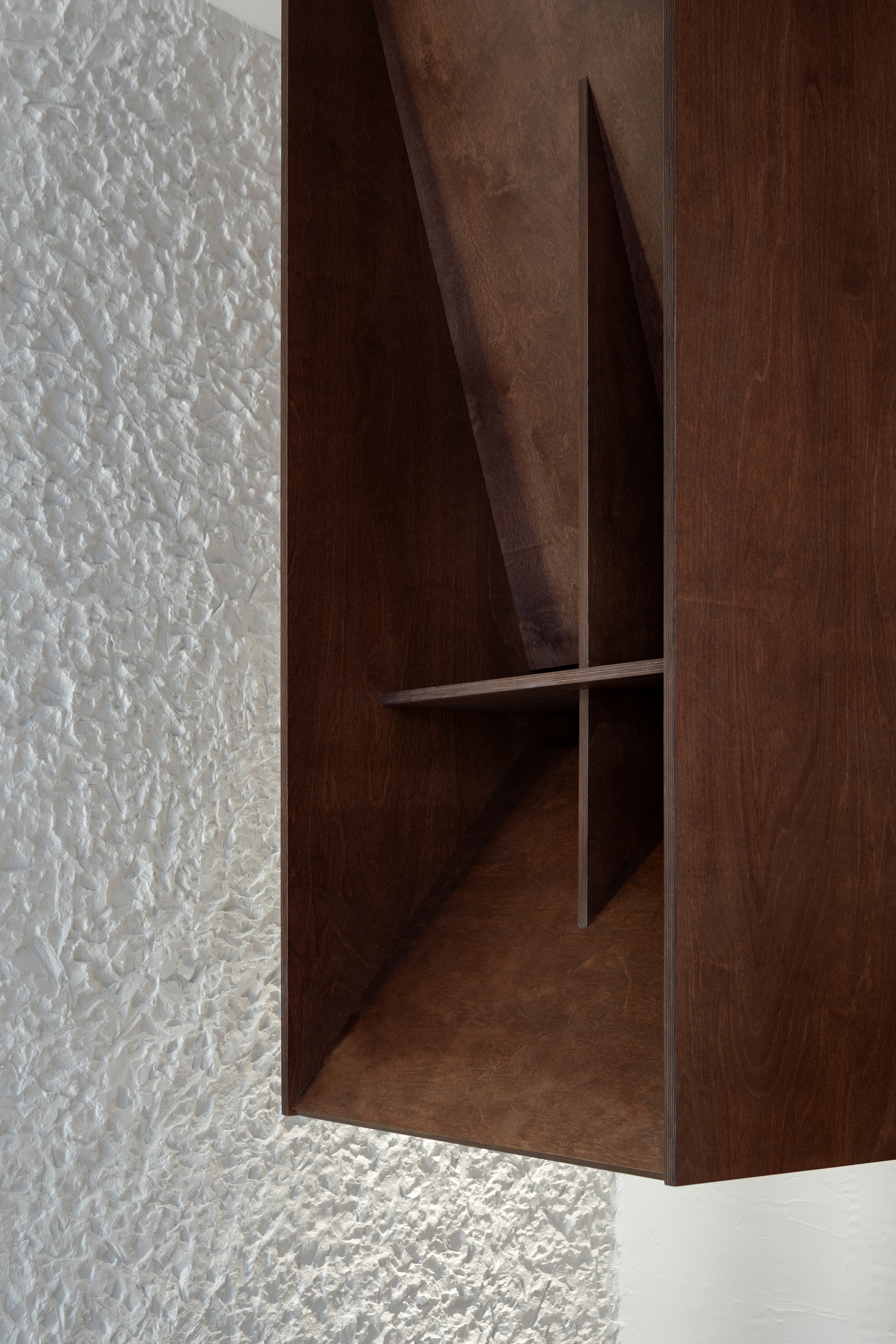 Triangular opening in a suspended walnut cabinet revealing interior shelving against textured plaster