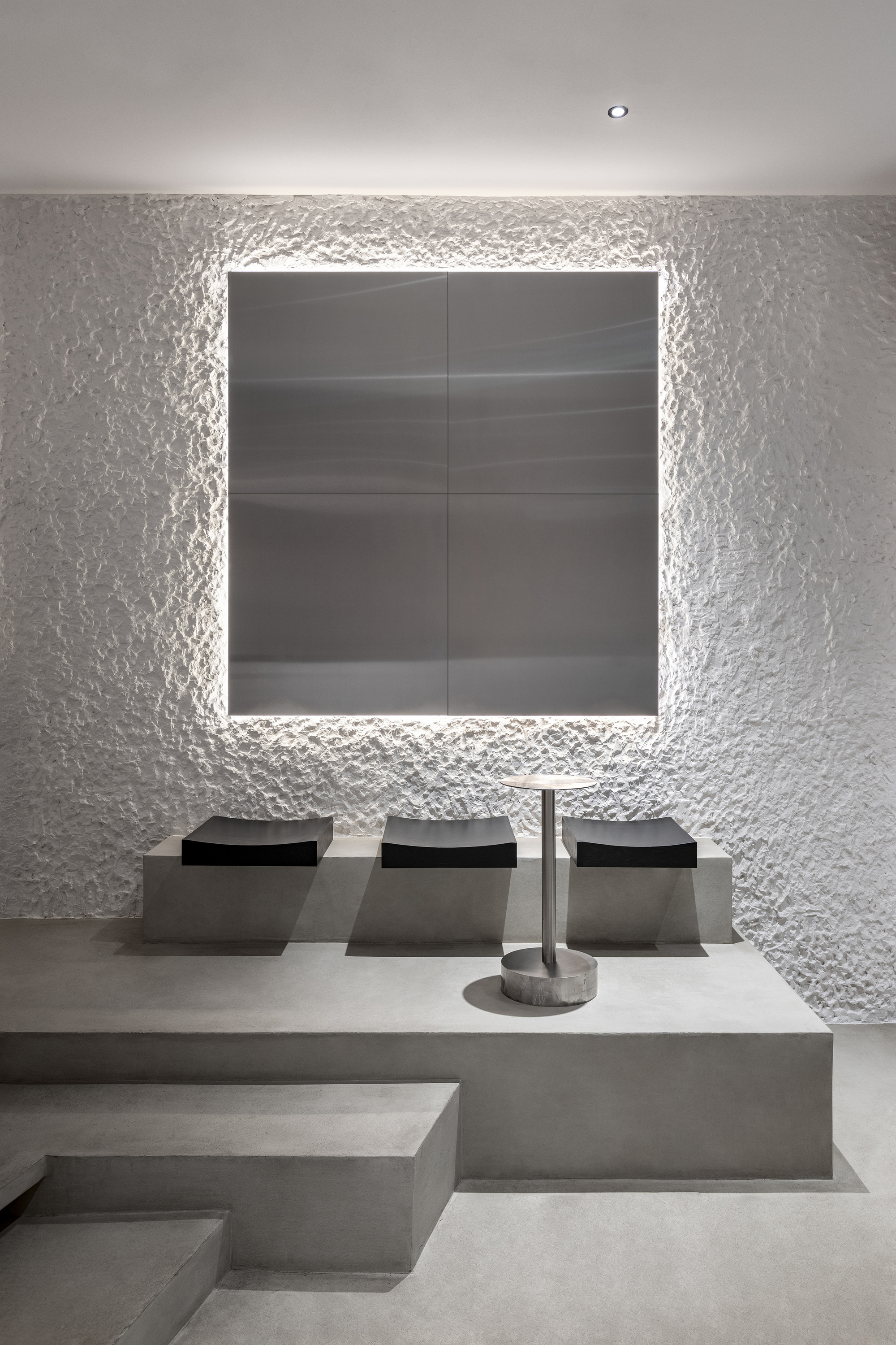 Stepped concrete seating platform facing backlit dark panel framed by rough white plastered walls