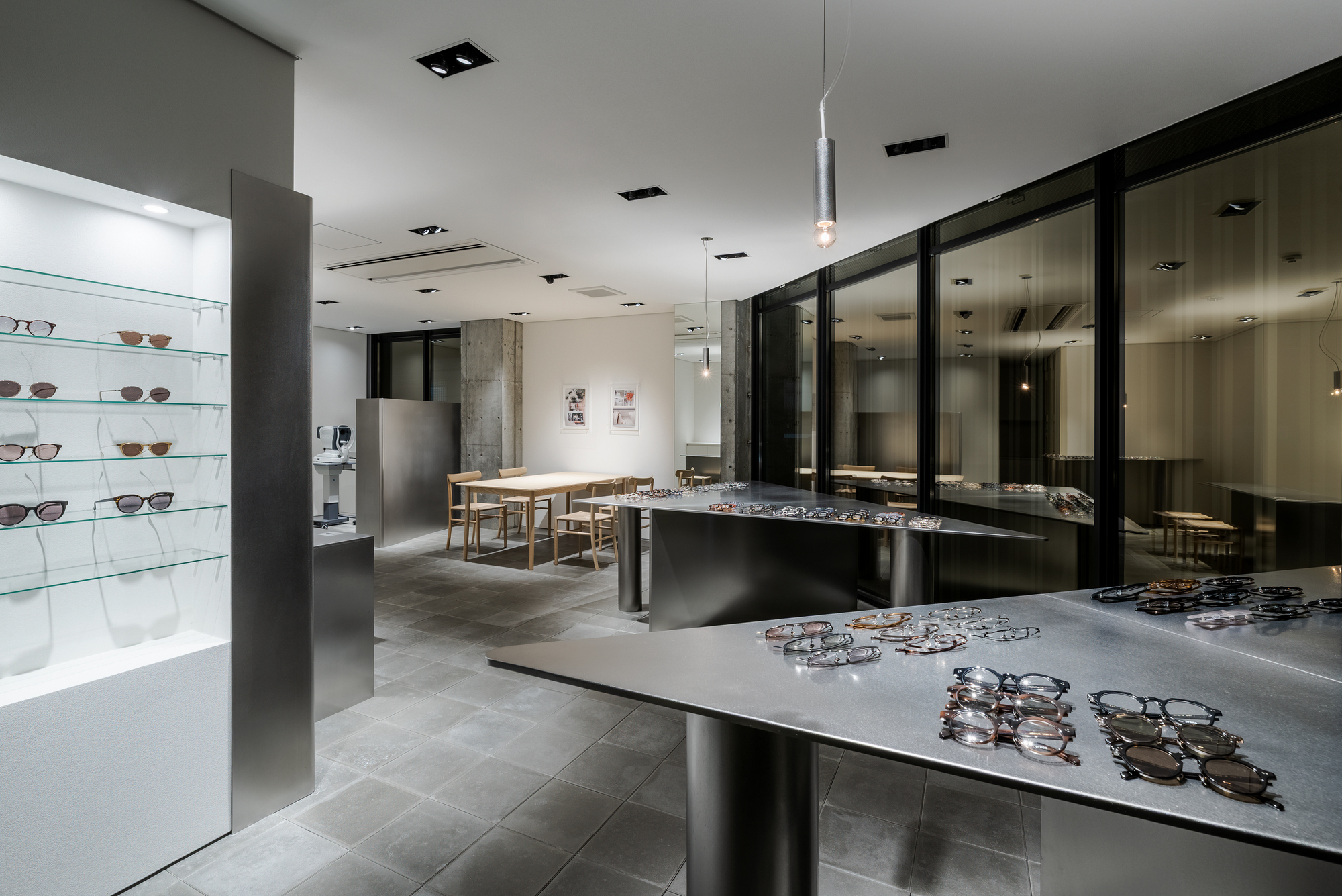 View across grey tiled floor showing eyeglass displays on metal tables and backlit shelving