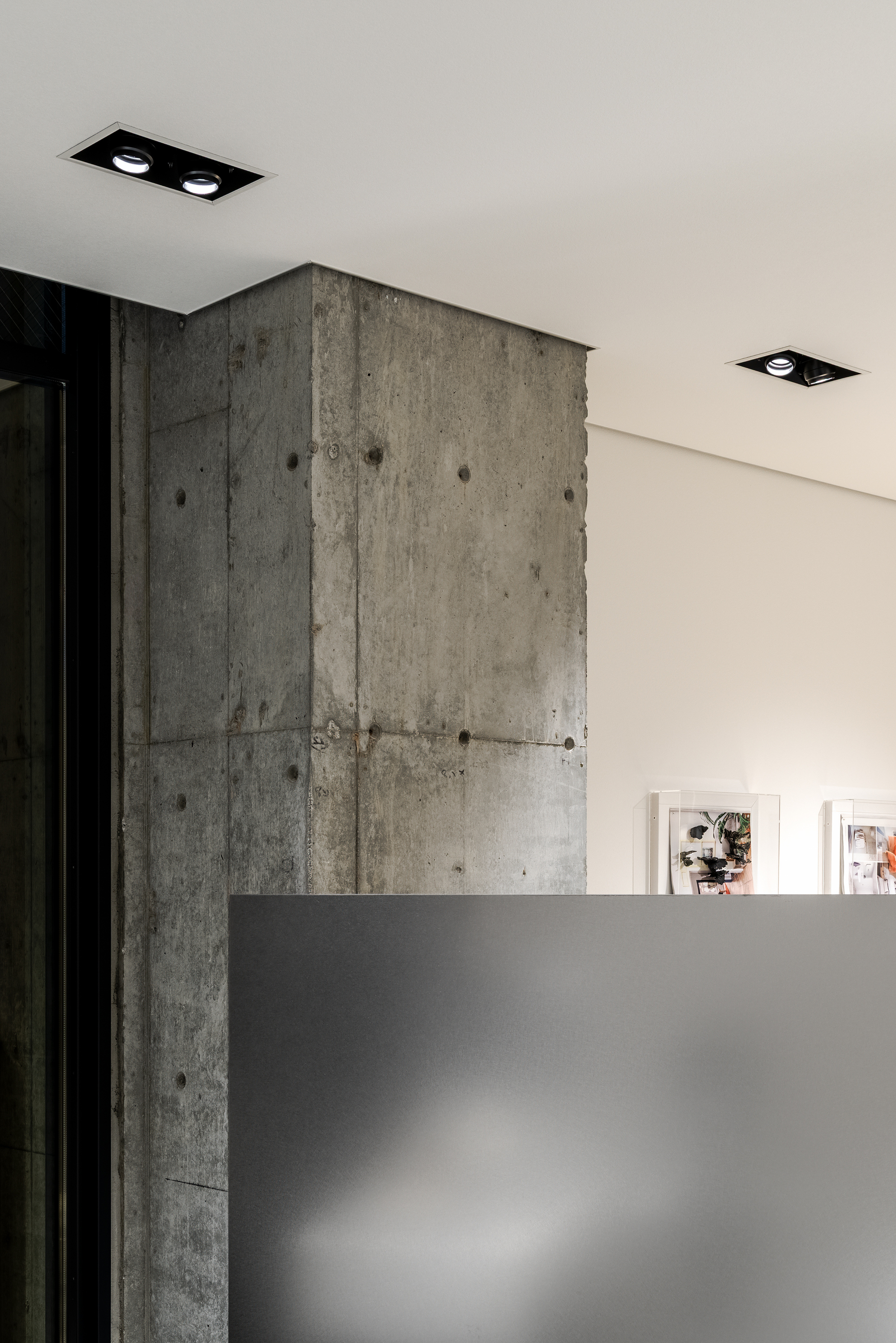 Board-formed concrete column rising behind a grey partition wall with recessed ceiling fixtures above