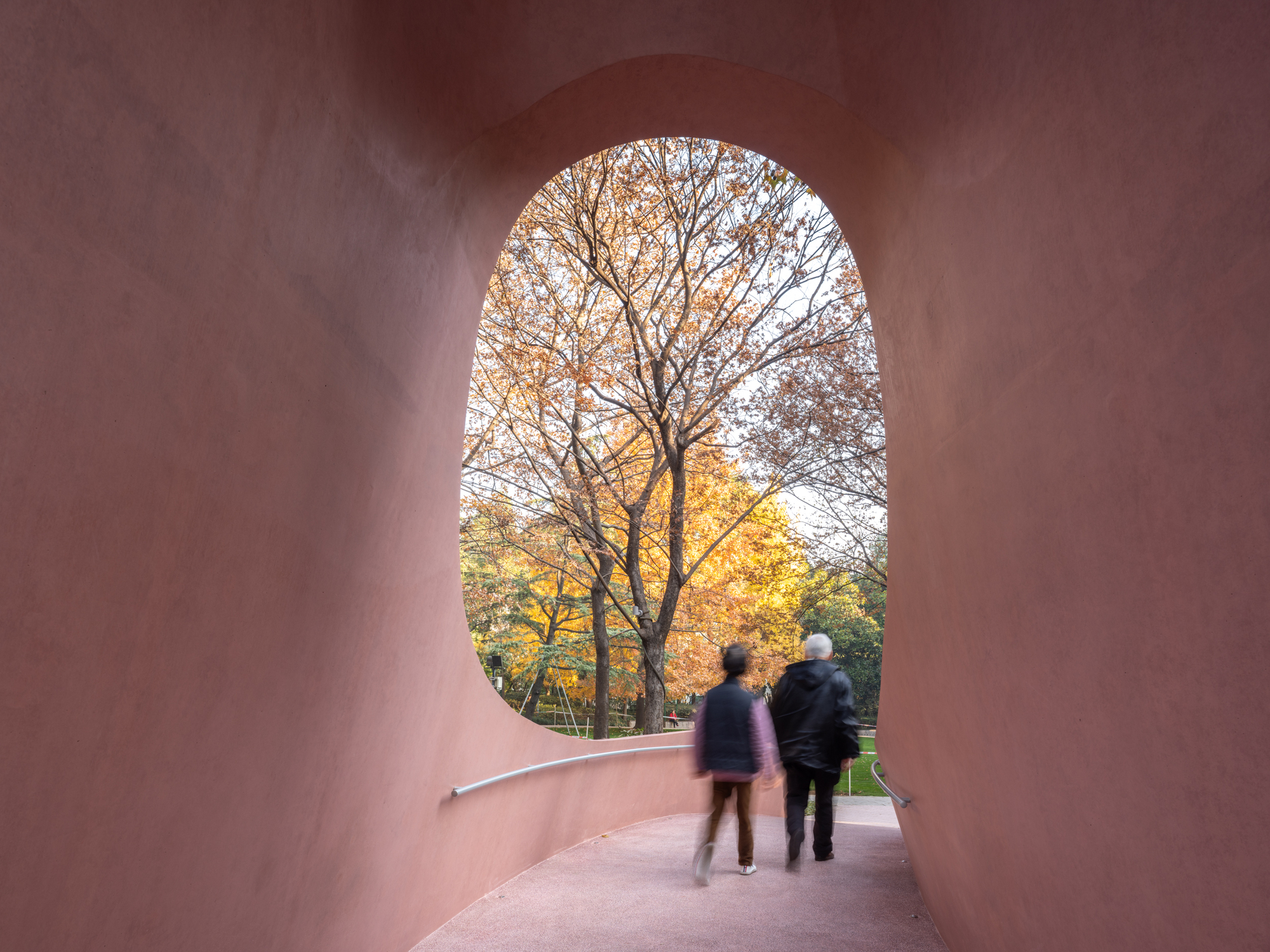 Interior tunnel view through arched pink concrete passage framing two visitors and autumn foliage beyond