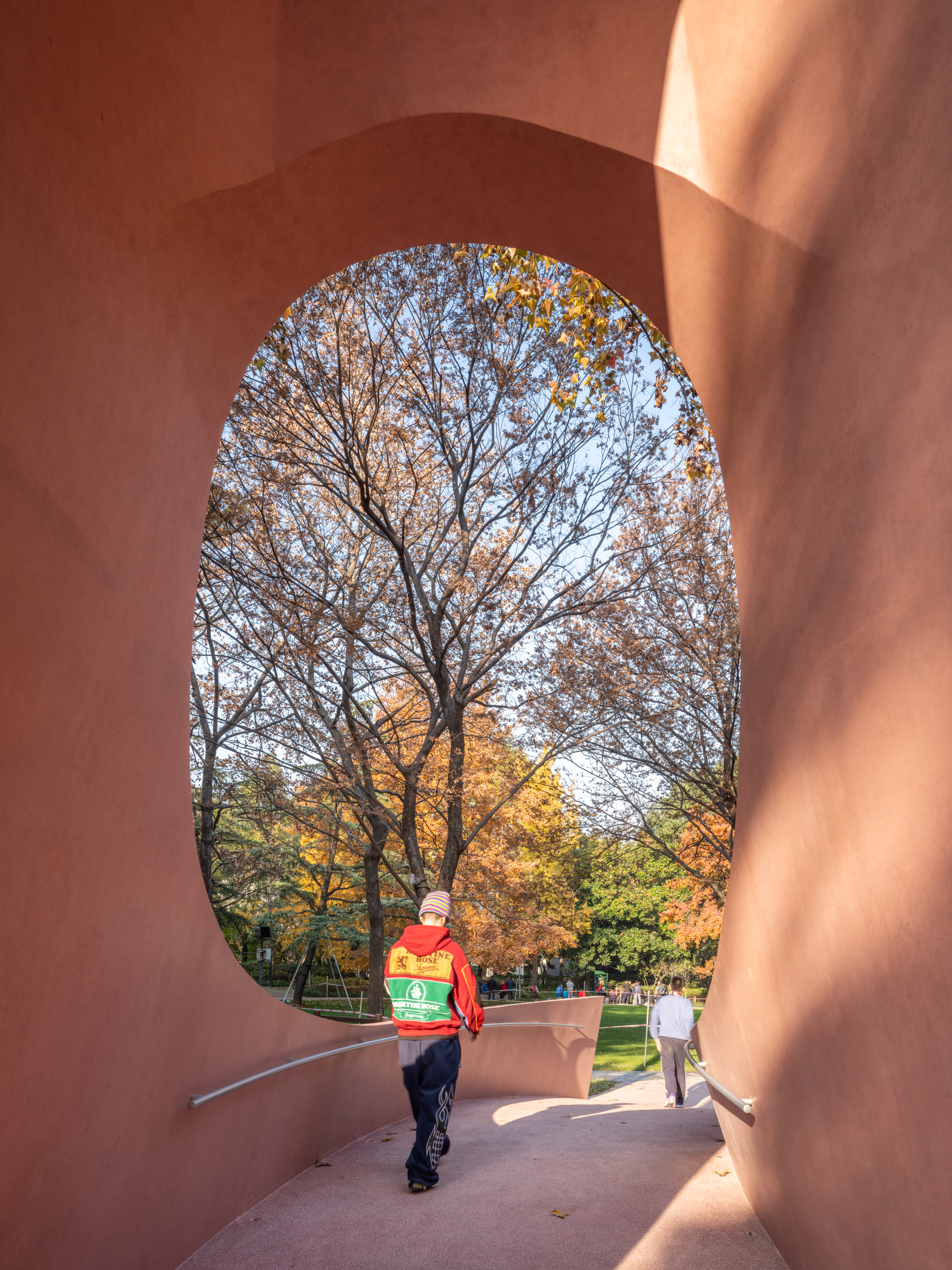 Visitor in red jacket walking through arched pink passage framing autumn landscape and park visitors