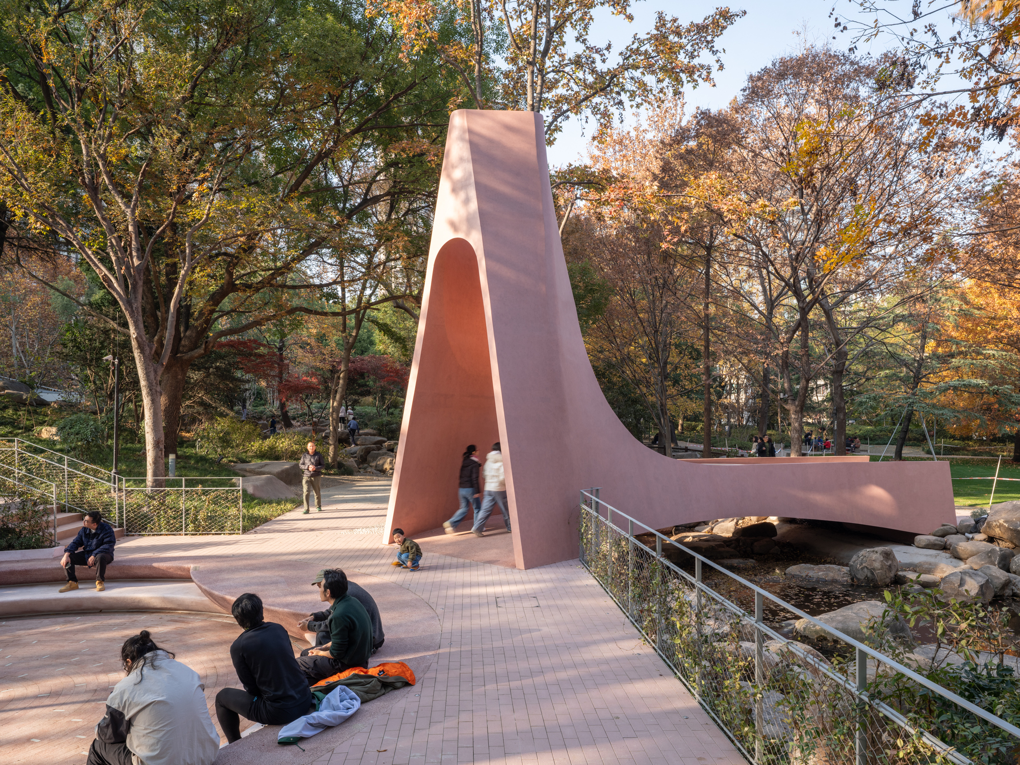 Sculptural pavilion with arched opening spanning a park pathway under dappled autumn sunlight