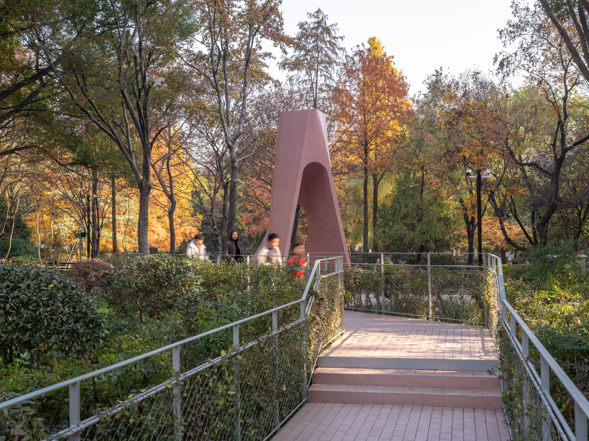 Mesh-railed pedestrian bridge leading to the pink arch structure at dusk with backlit trees