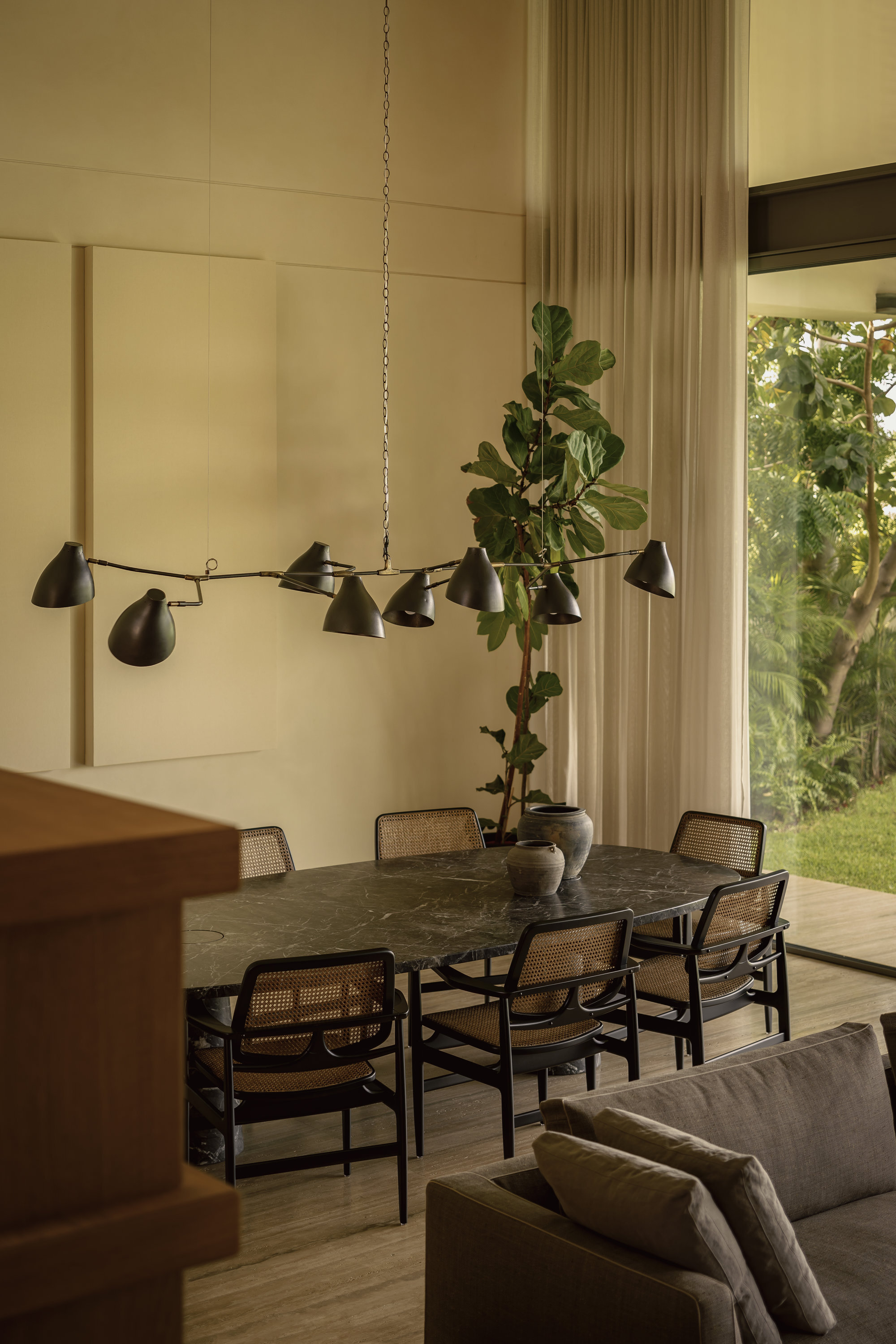 Dining area with chain-suspended chandelier above dark table overlooking garden through sheer curtains