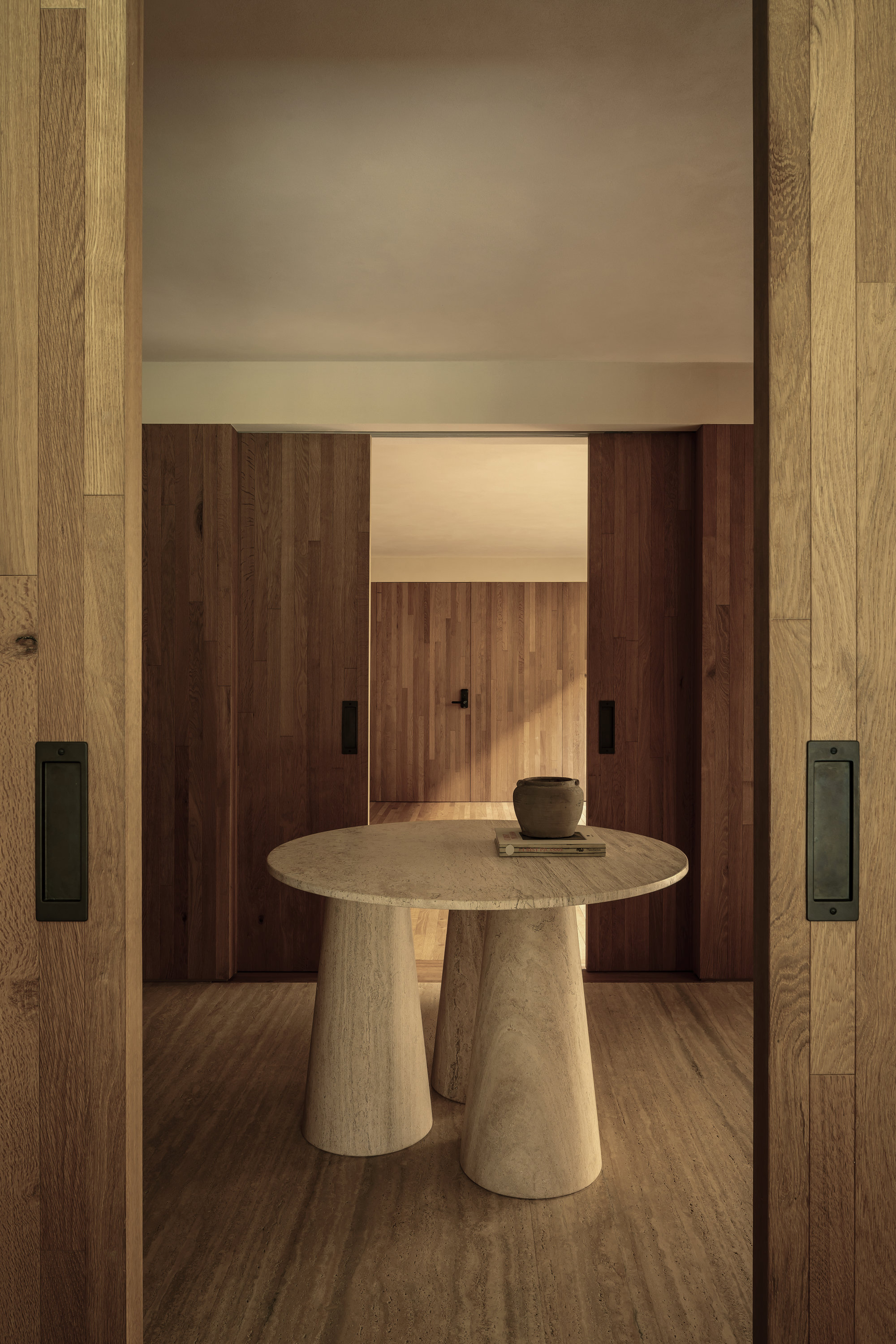 View through sliding oak doors into a vestibule with a stone pedestal table under warm lighting
