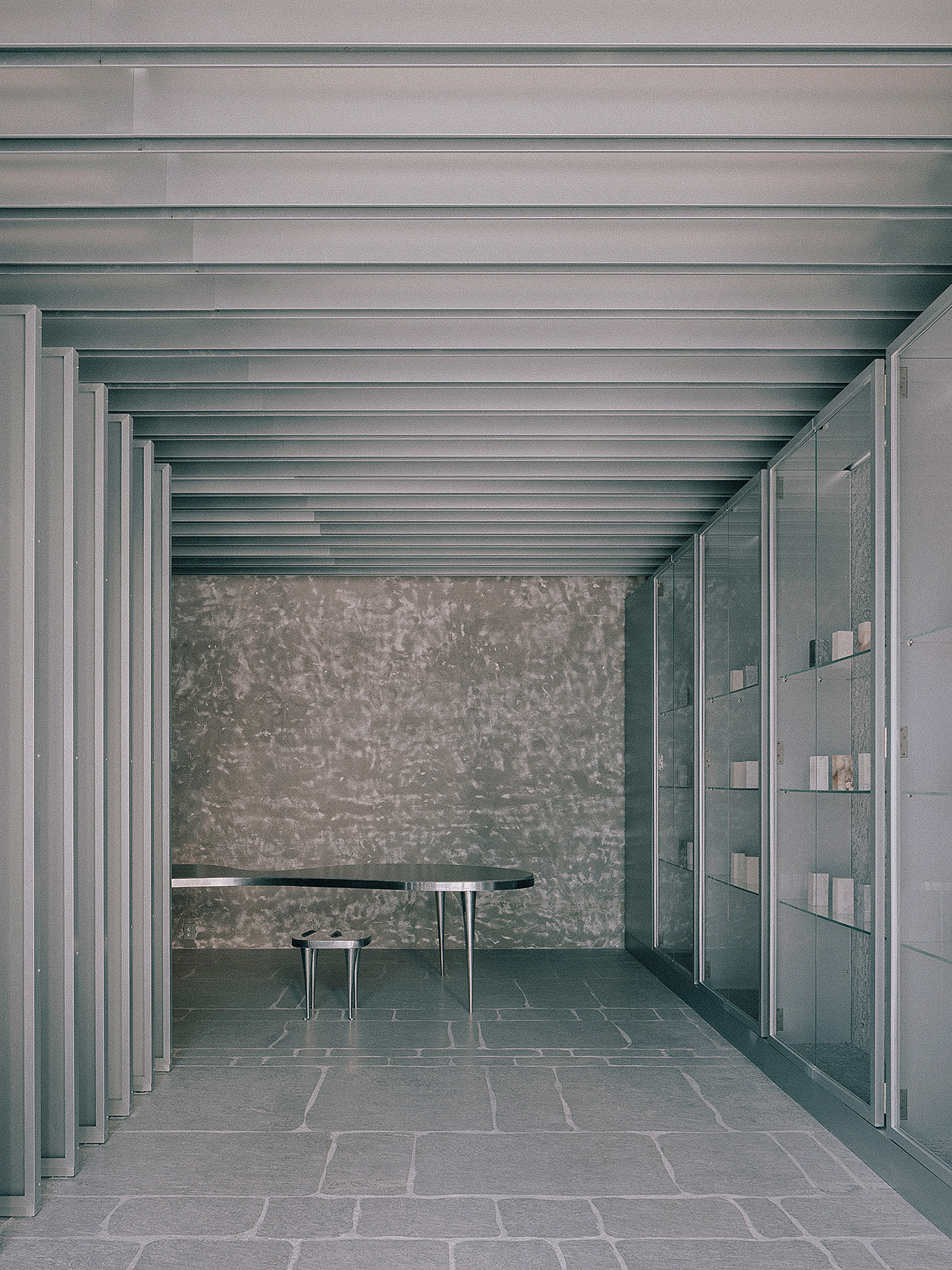 Interior colonnade with exposed metal beams, textured stone wall, dark table and floor-to-ceiling glazed partition