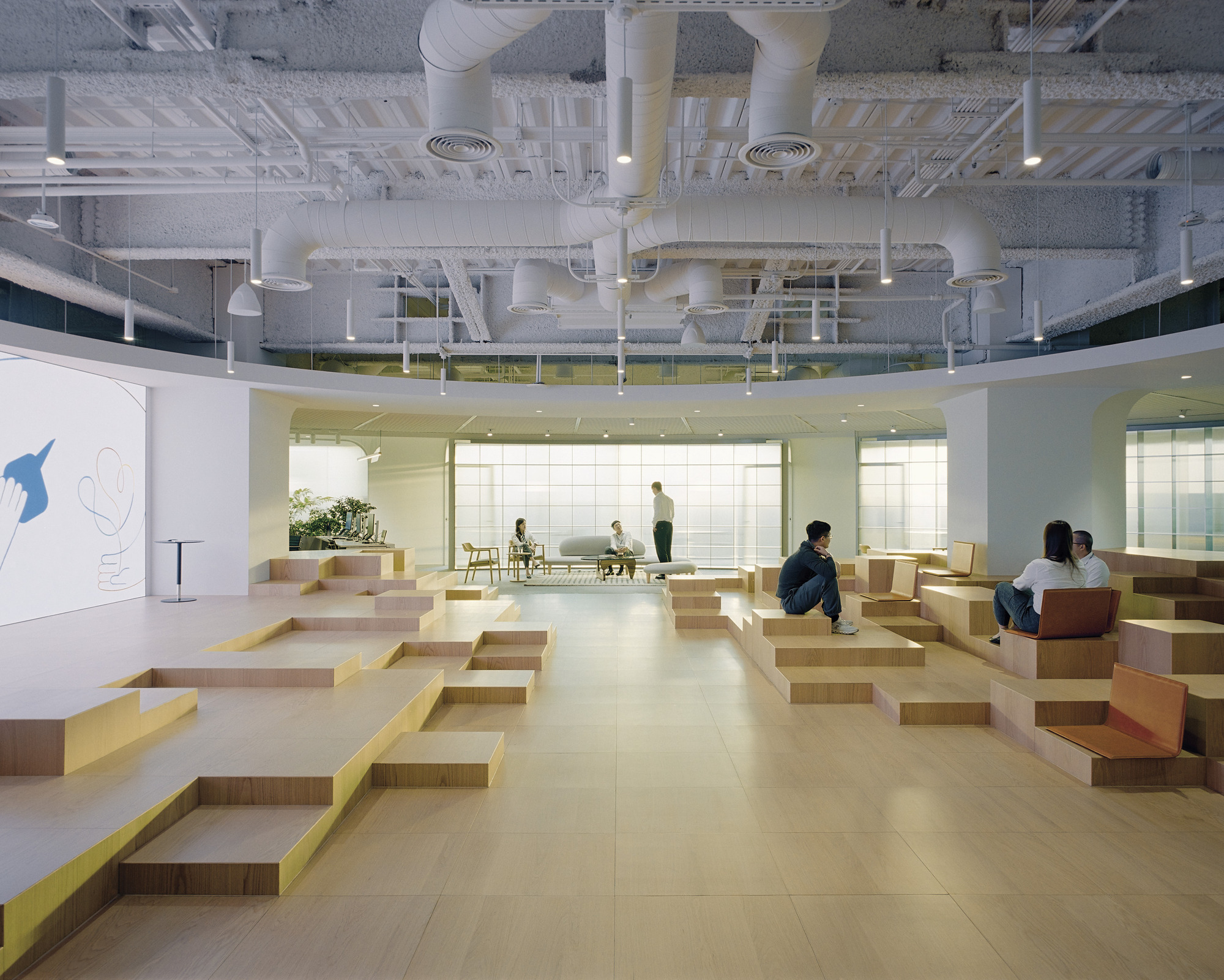 Tiered plywood seating platforms in an open office atrium with exposed ductwork and mezzanine above