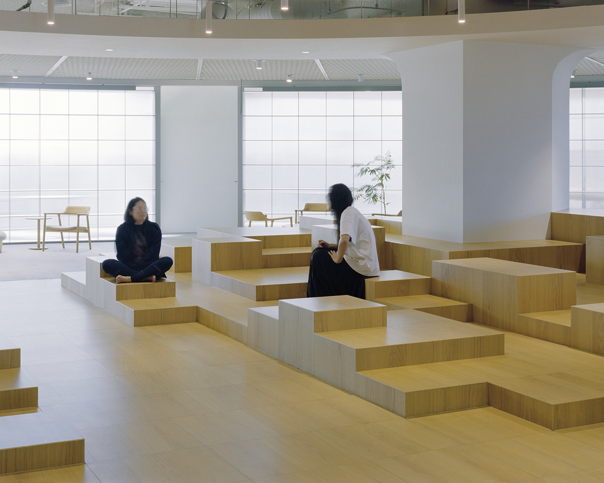 Two visitors seated on geometric timber block seating near gridded translucent glass facade