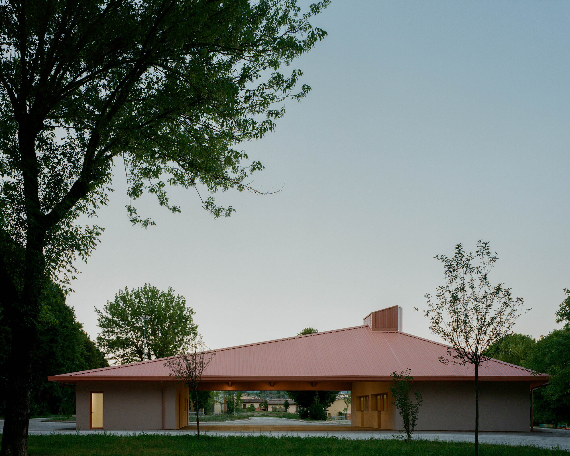 Pavilion with broad hipped metal roof and open-air loggia framed by young trees at dusk