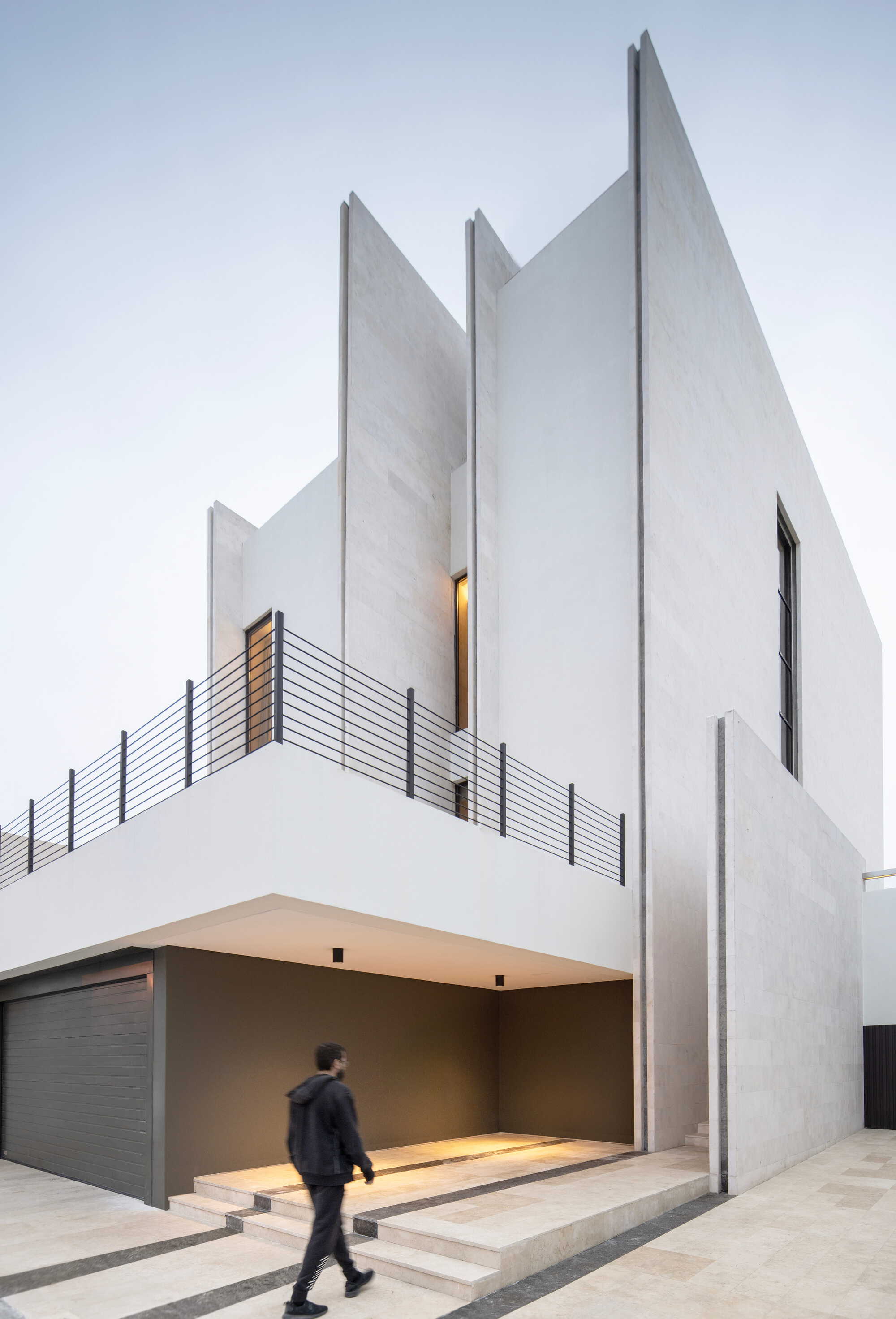 White concrete facade with jagged peaks and a person walking through the covered garage entry at dusk