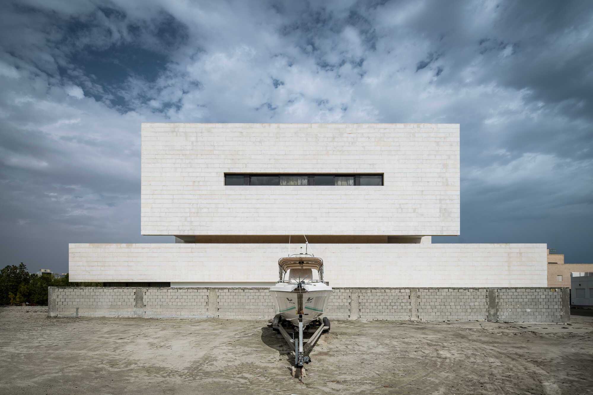 Layered white limestone facade with horizontal slot window and boat centered in forecourt under stormy clouds
