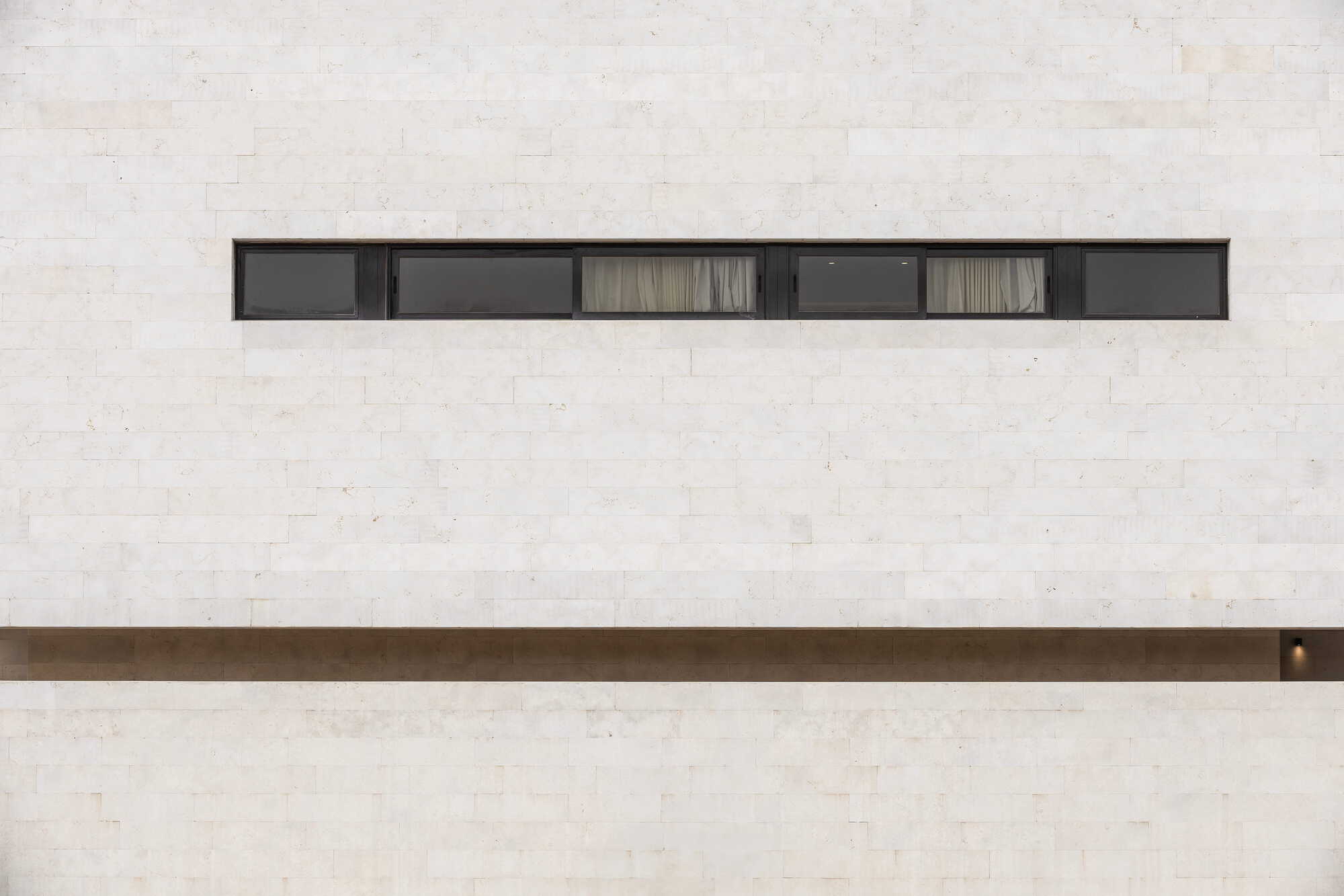 Horizontal ribbon window with six panels set into a pale travertine facade with bronze detailing below