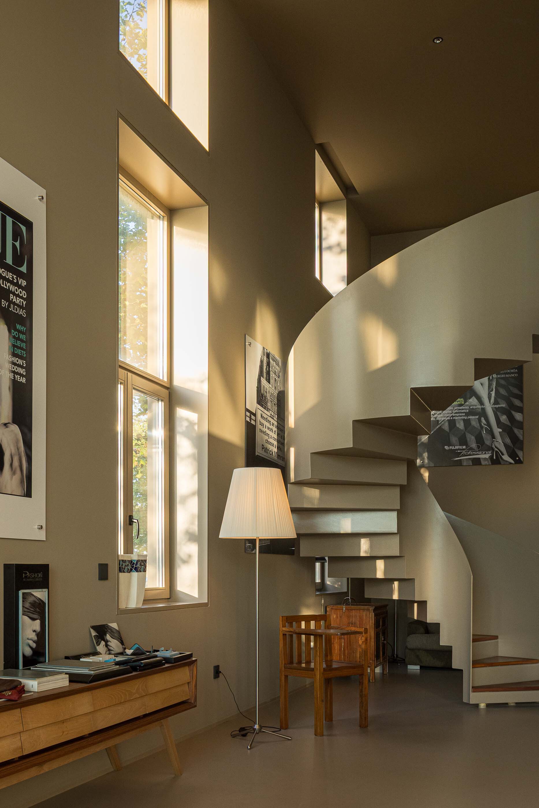 Curved floating staircase bathed in sunlight from tall windows beside framed posters