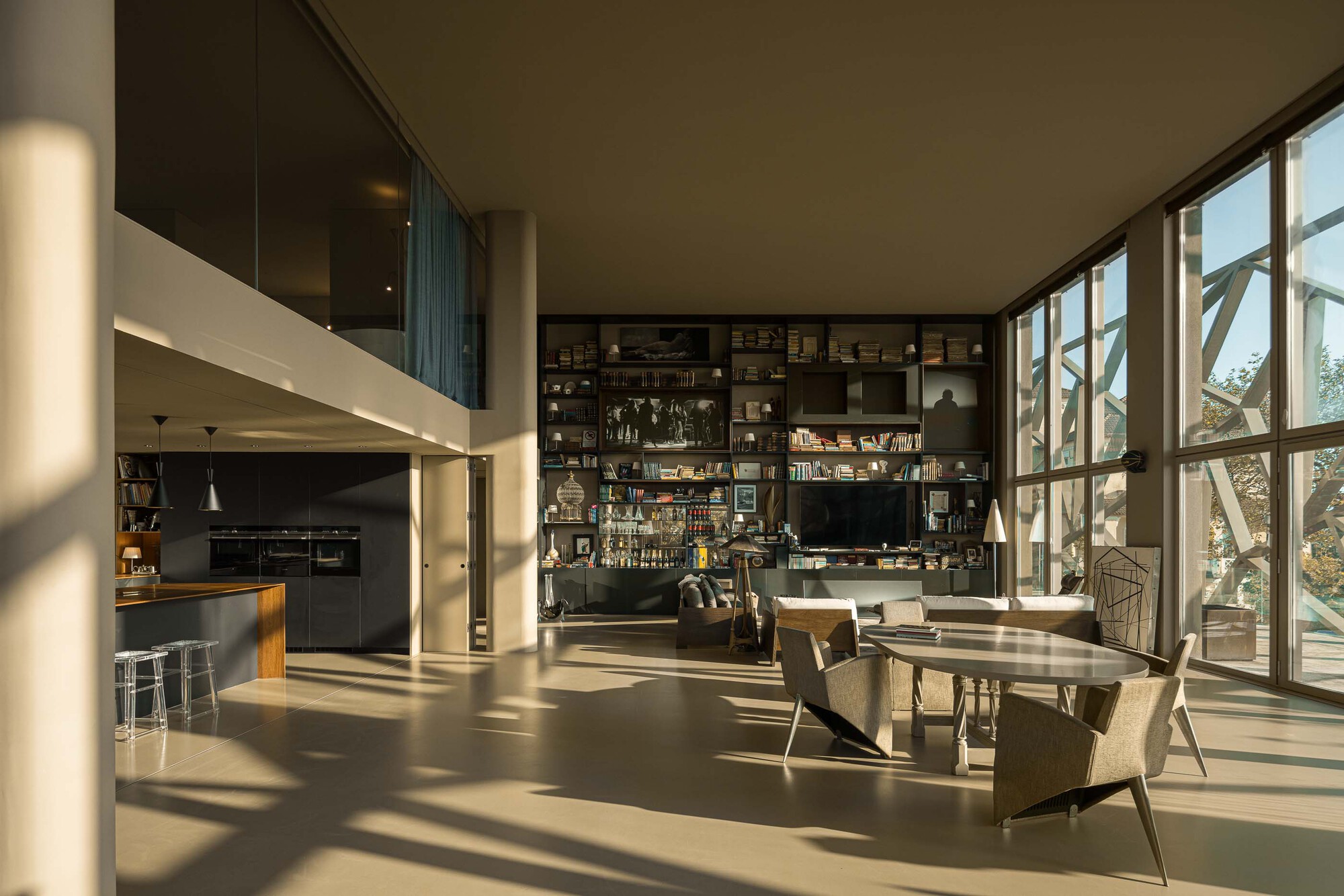 Double-height living space with floor-to-ceiling windows, open kitchen, and floating shelving wall in afternoon sunlight