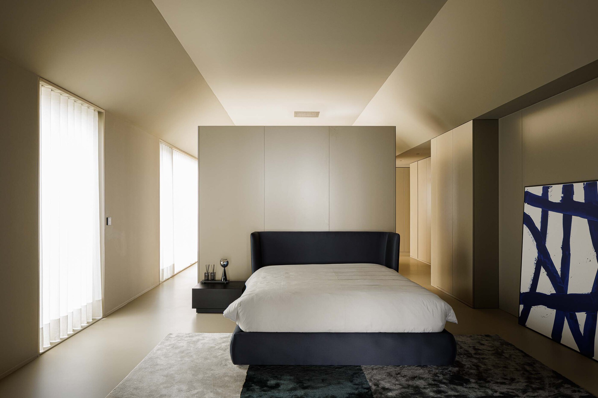 Bedroom with black upholstered bed, backlit storage wall, and filtered light from sheer curtains