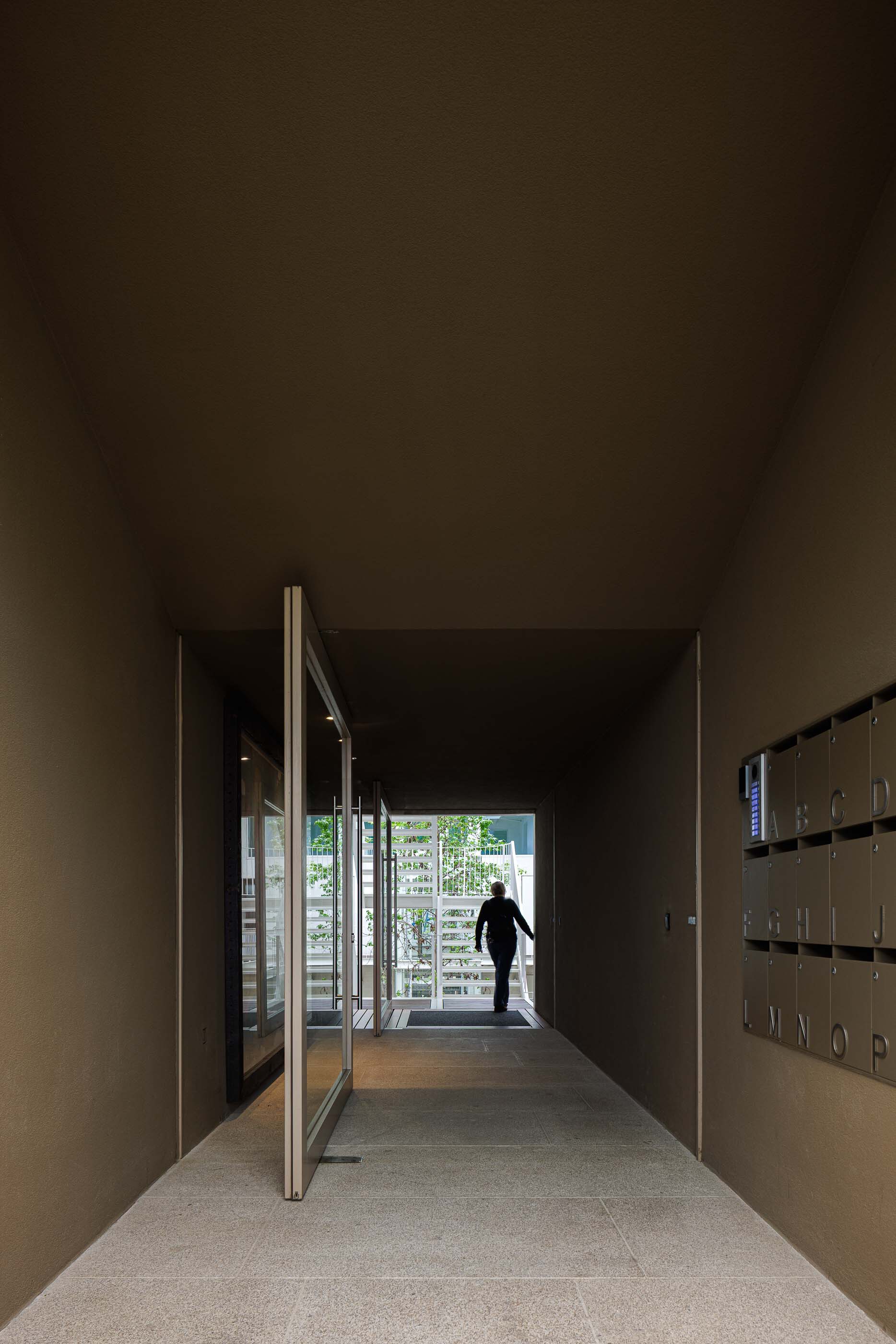 Entry corridor with pivoting glass door and terrazzo flooring leading to a figure in backlit courtyard