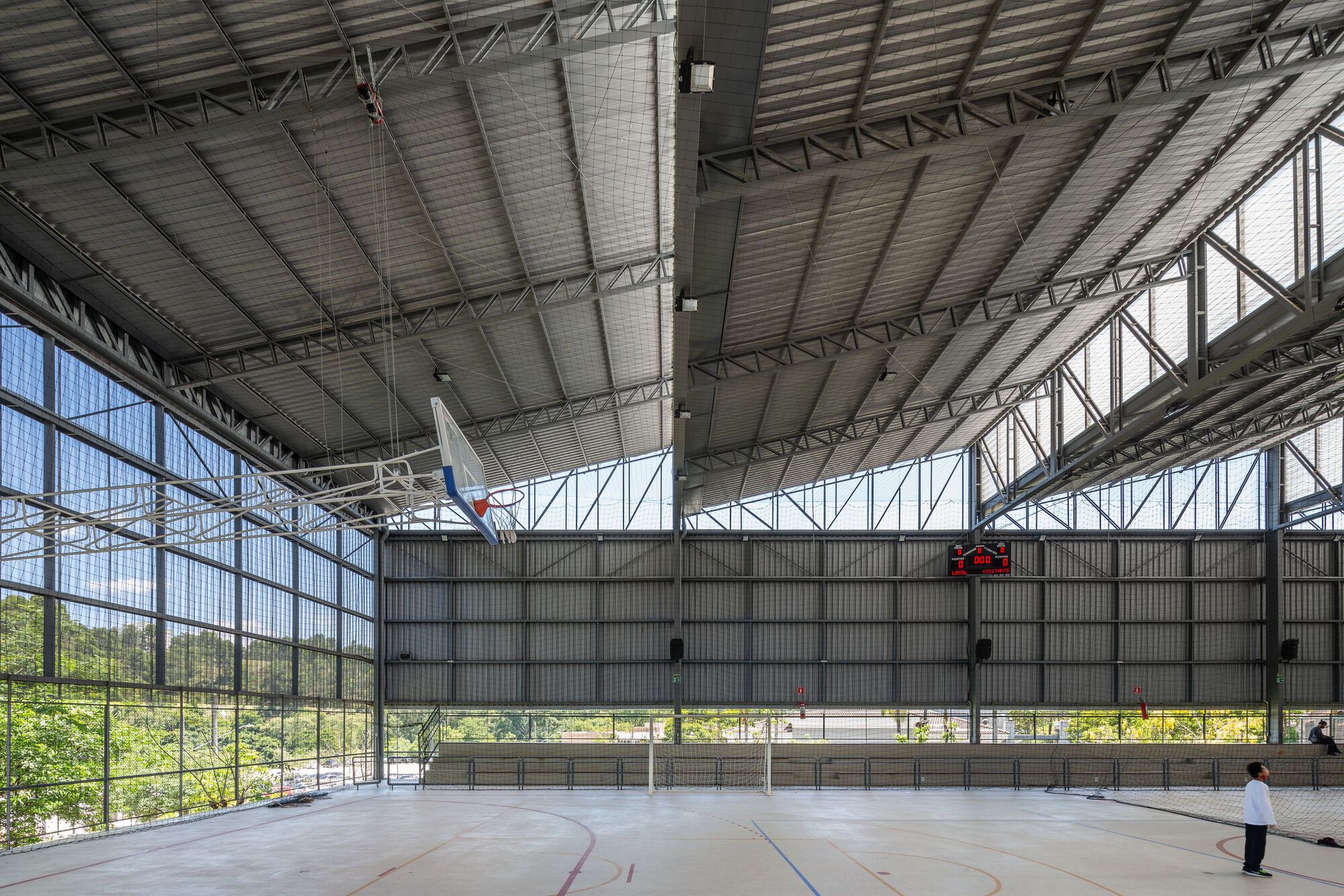 Interior sports court with corrugated metal ceiling and exposed steel trusses overlooking greenery through glazed walls