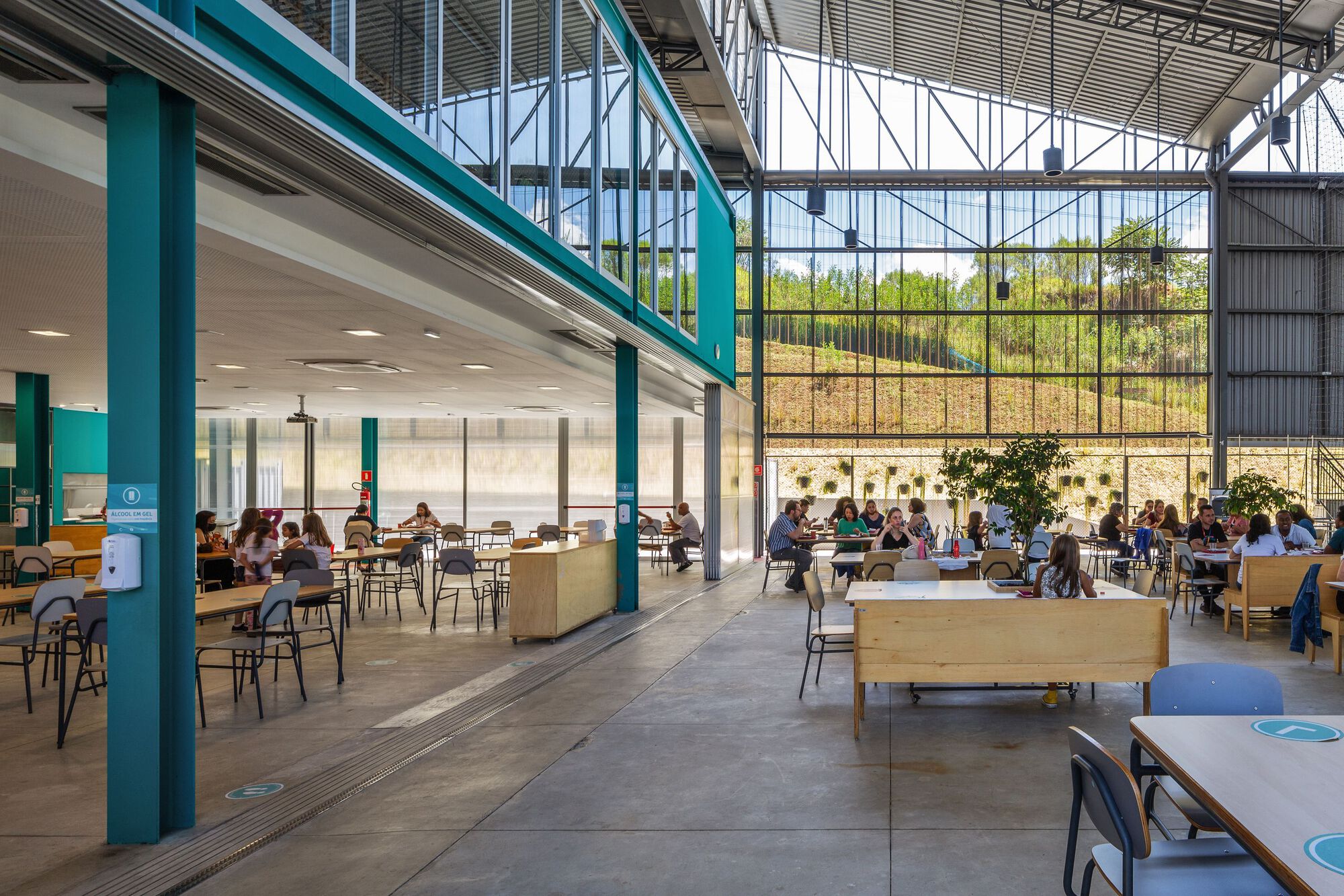 Cafeteria space framed by teal steel columns with light wood benches and people gathering beneath exposed trusses