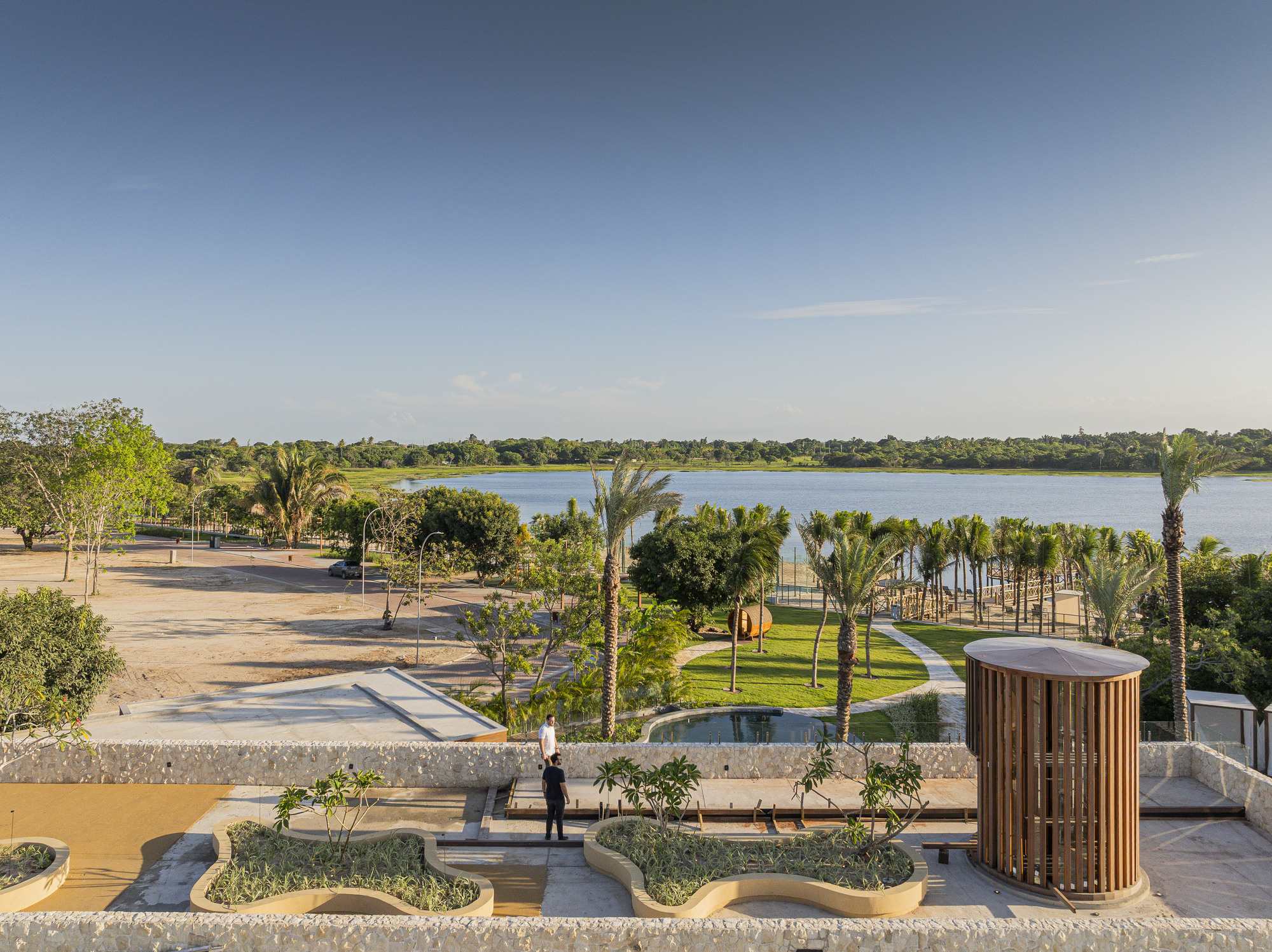 Tiered concrete plaza with planted islands and cylindrical timber tower overlooking a distant lake