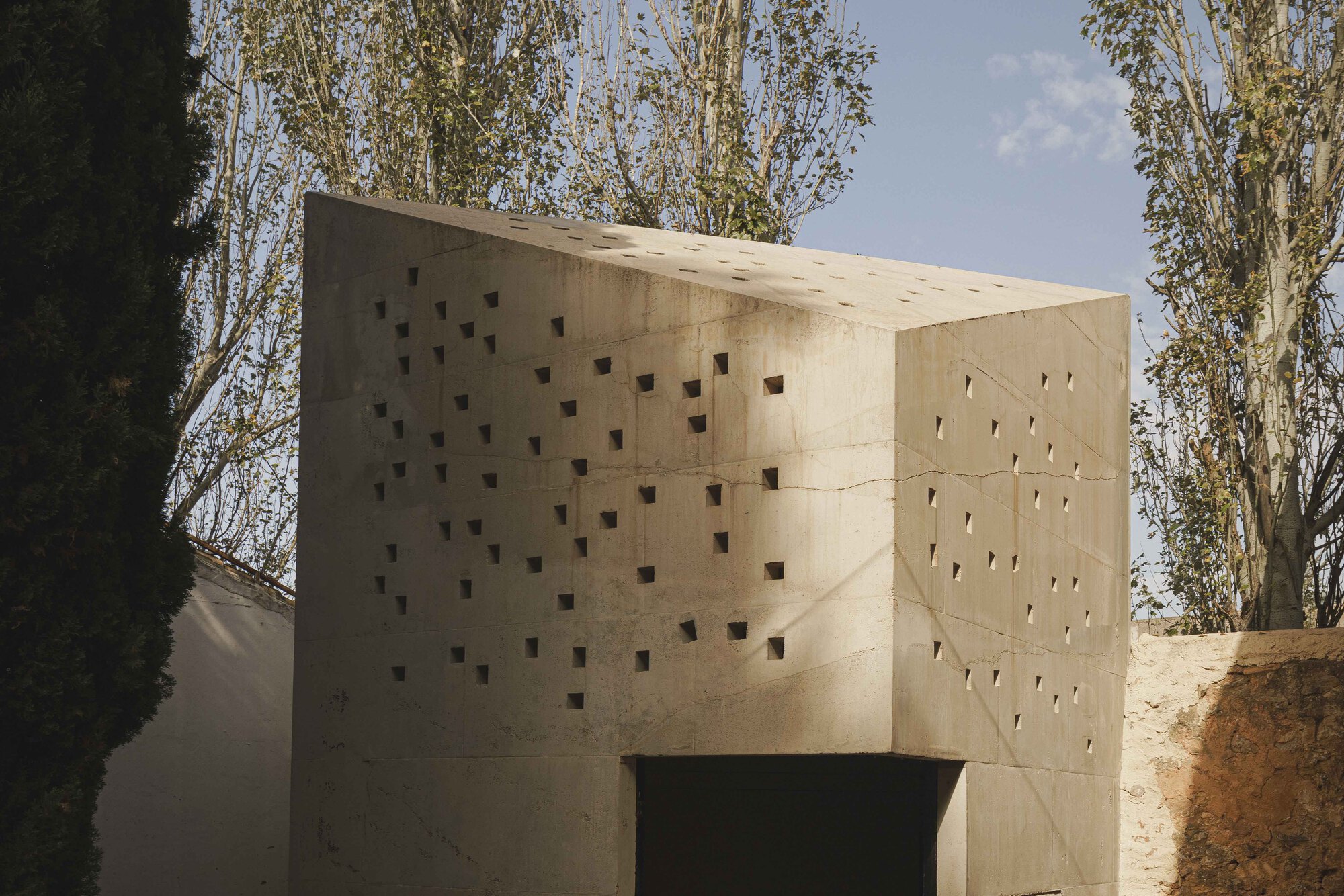 Concrete volume with grid of square perforations set among deciduous trees in afternoon light