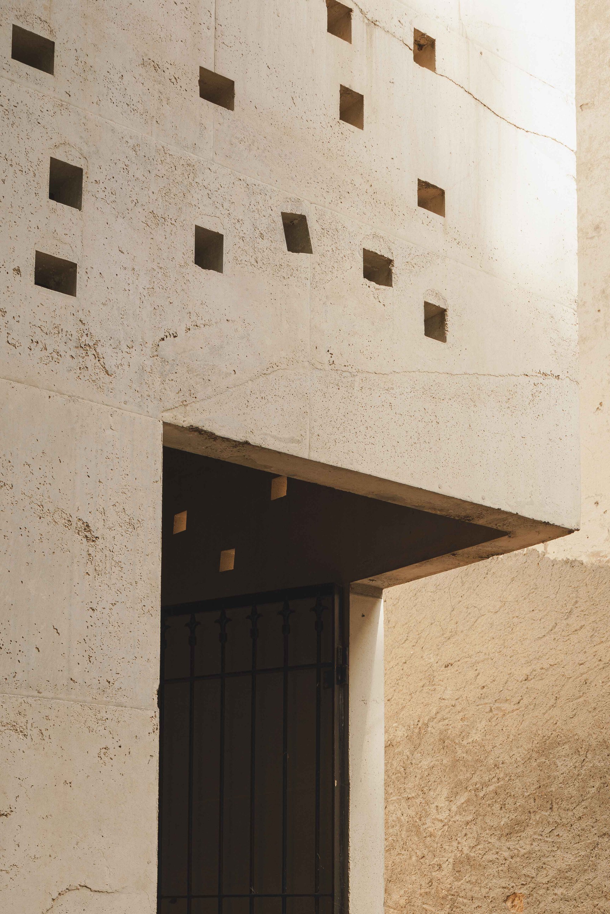 Triangular canopy over a timber door with square openings dotting the concrete wall above