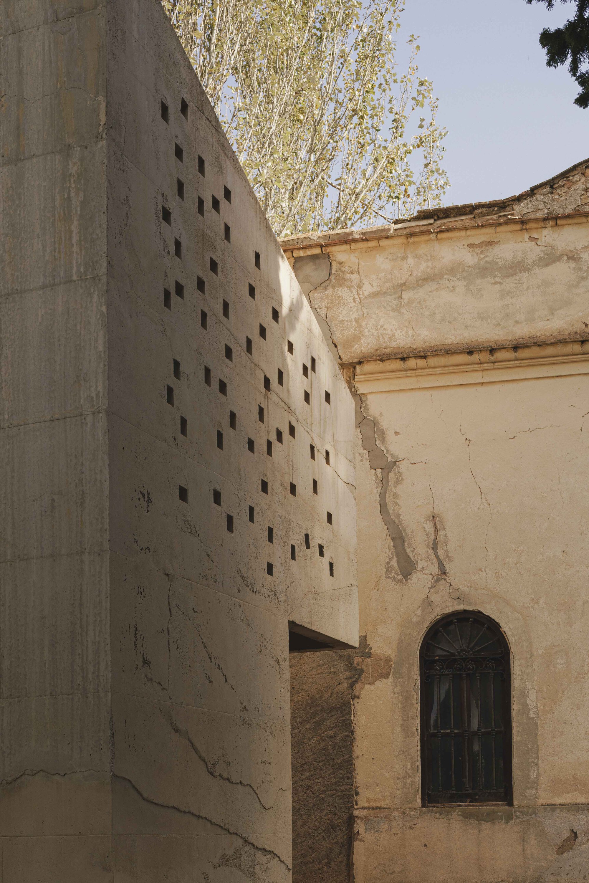 Perforated concrete wall rising beside weathered masonry facade with arched door and tree beyond