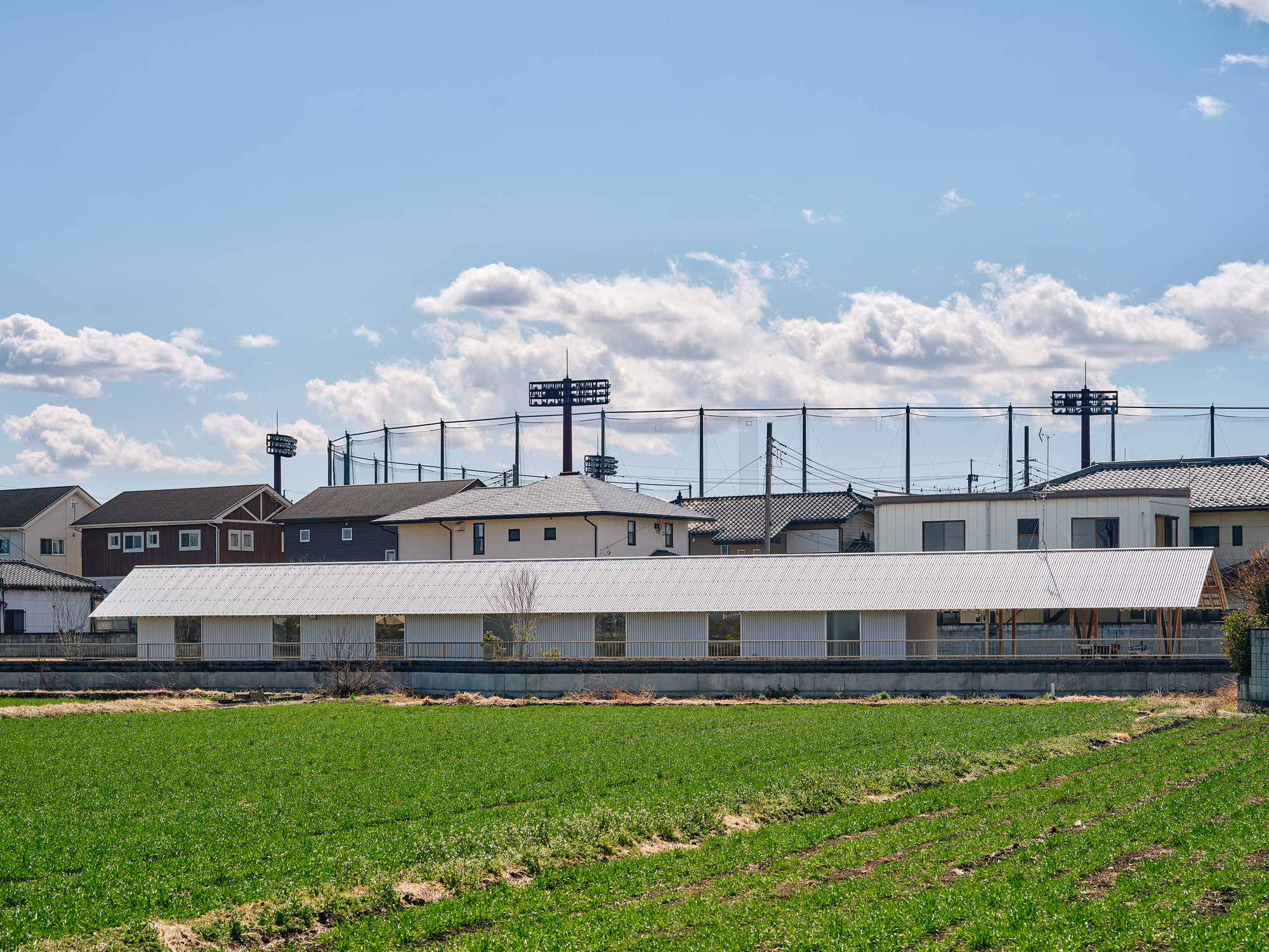 Low horizontal volume with white corrugated metal roof beyond green agricultural field and suburban houses