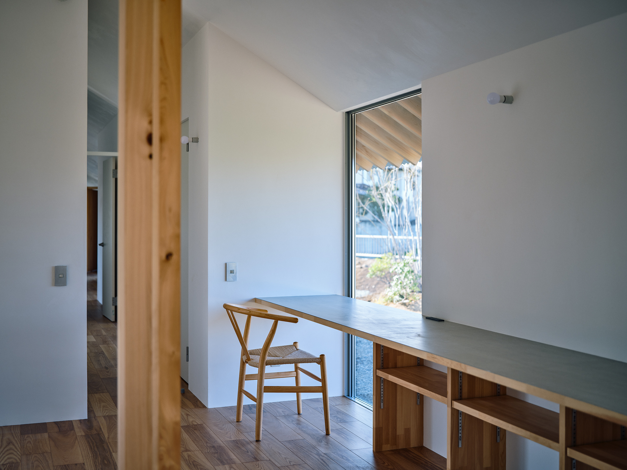 Built-in desk with timber shelving below a tall window framing views of shrubs and trees