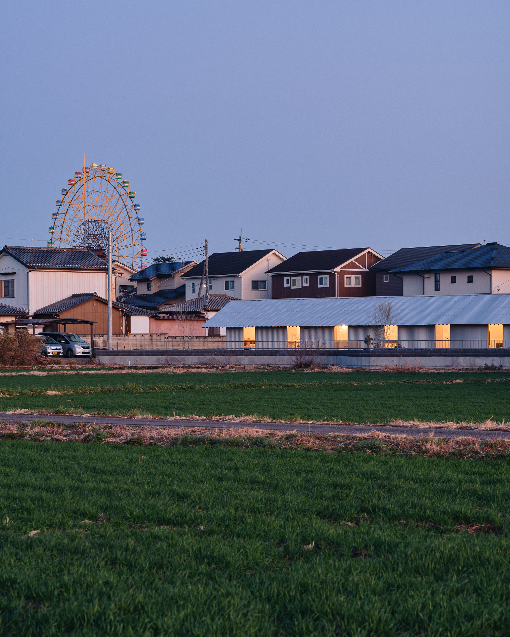 Long facade with evenly spaced illuminated openings viewed across a field at dusk with a ferris wheel behind