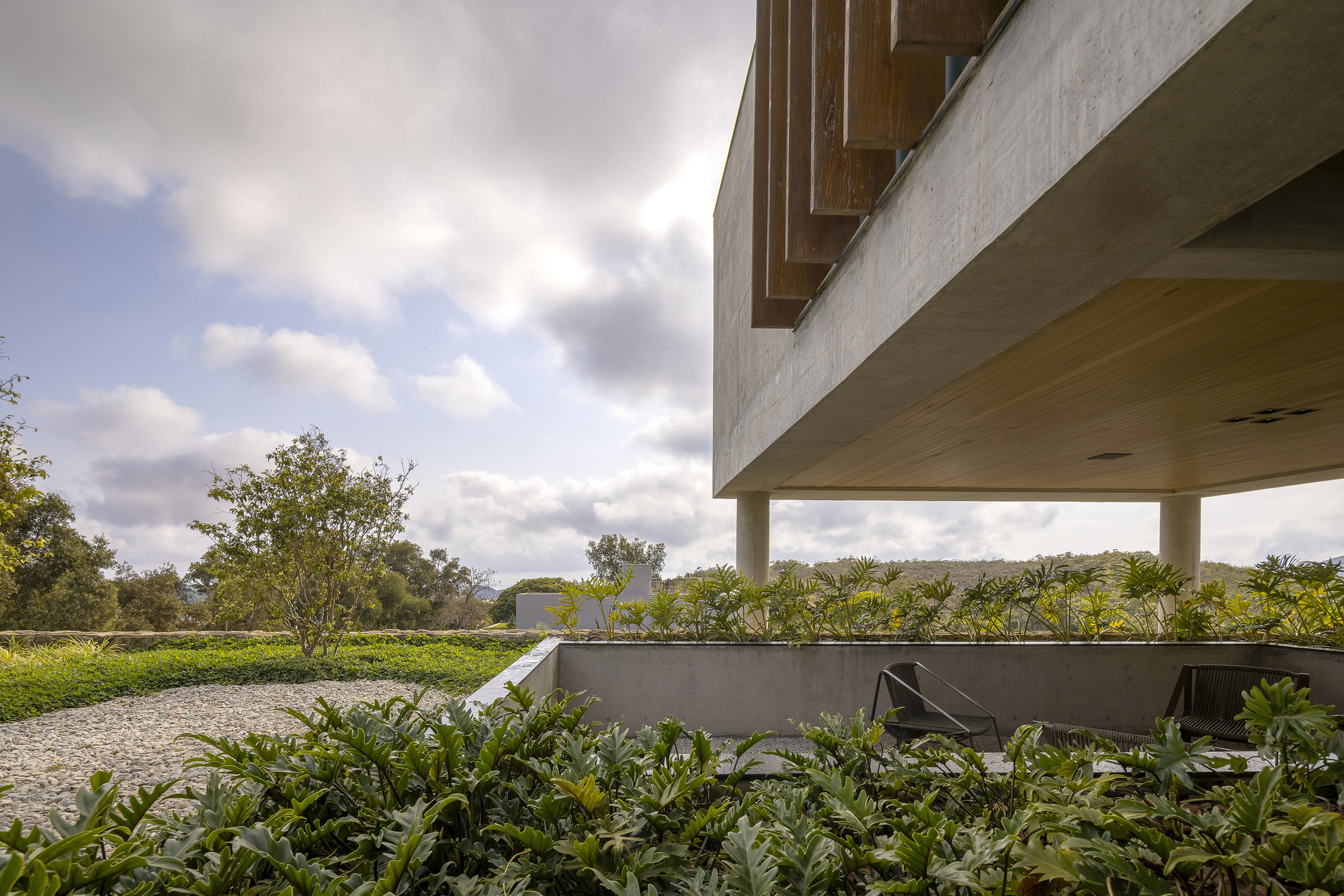 Cantilevered concrete volume above a planted terrace with gravel path and cloudy sky