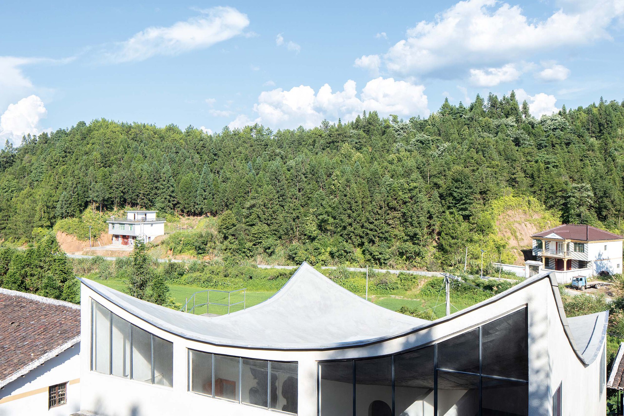 Curving white roof canopy above floor to ceiling glazing overlooking the valley under summer clouds