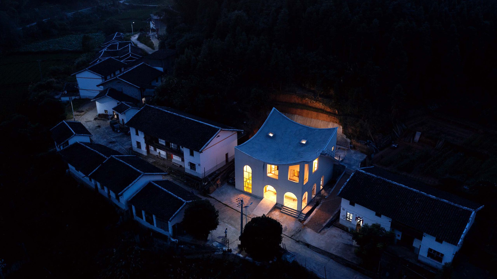 Aerial view at dusk showing the illuminated arched windows against the dark surrounding village rooftops