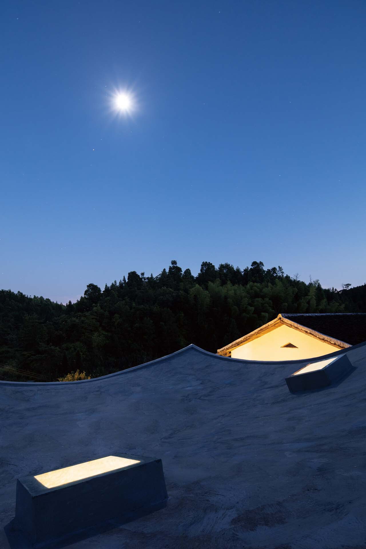 Rooftop terrace with curved parapets and a lit gable skylight under moonlight against a forested hillside