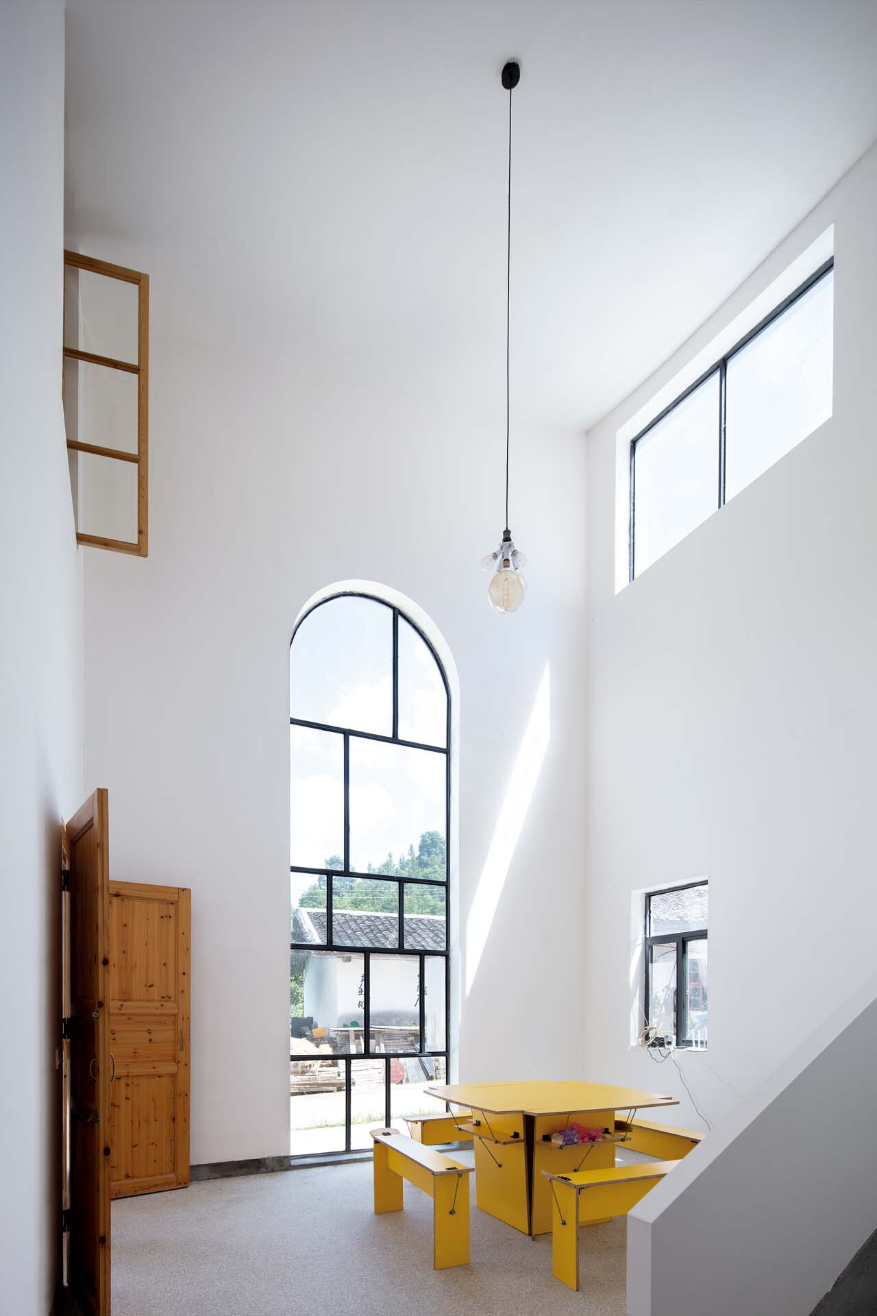 Tall arched steel window framing trees outside, with yellow table and pendant bulb in white interior
