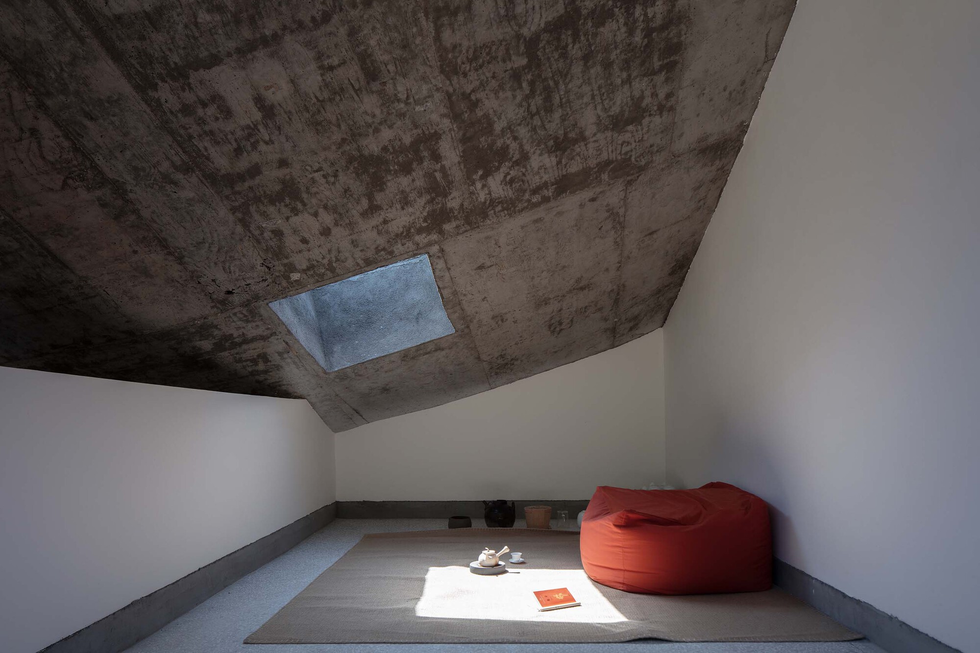 Minimalist room with board-formed concrete ceiling, rectangular skylight, raised platform, and orange bean bag
